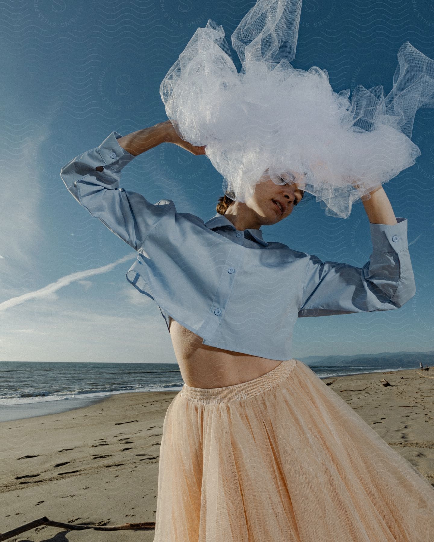 Woman in a blue shirt and peach skirt with a white veil over her head on the beach, clear blue sky in the background.