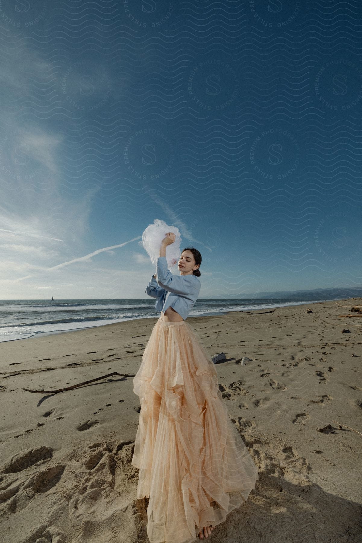 Woman in a peach skirt and blue top playing with a white fabric on a sandy beach with ocean and sky in the background.