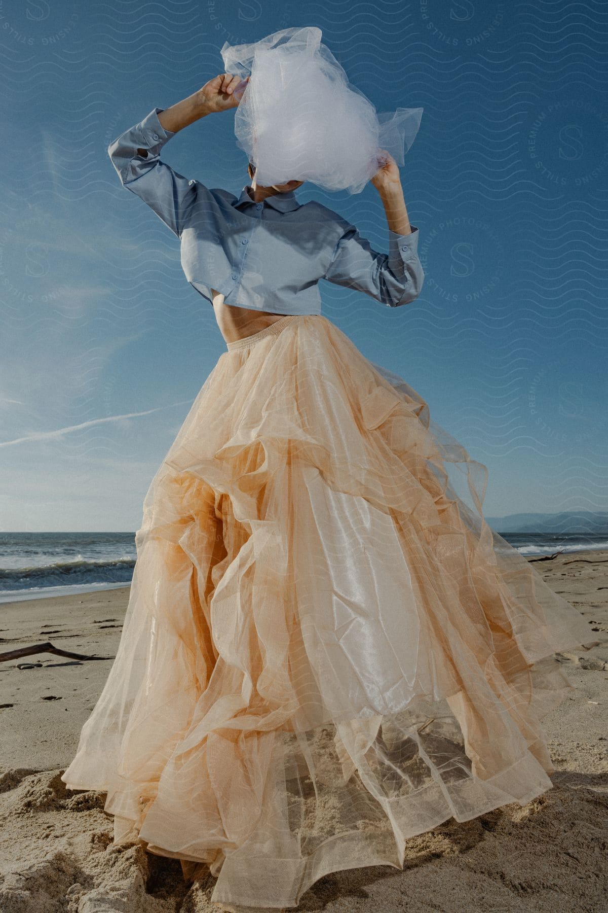 A woman in formal wear on the beach, with a veil wrapped around her head hiding her face