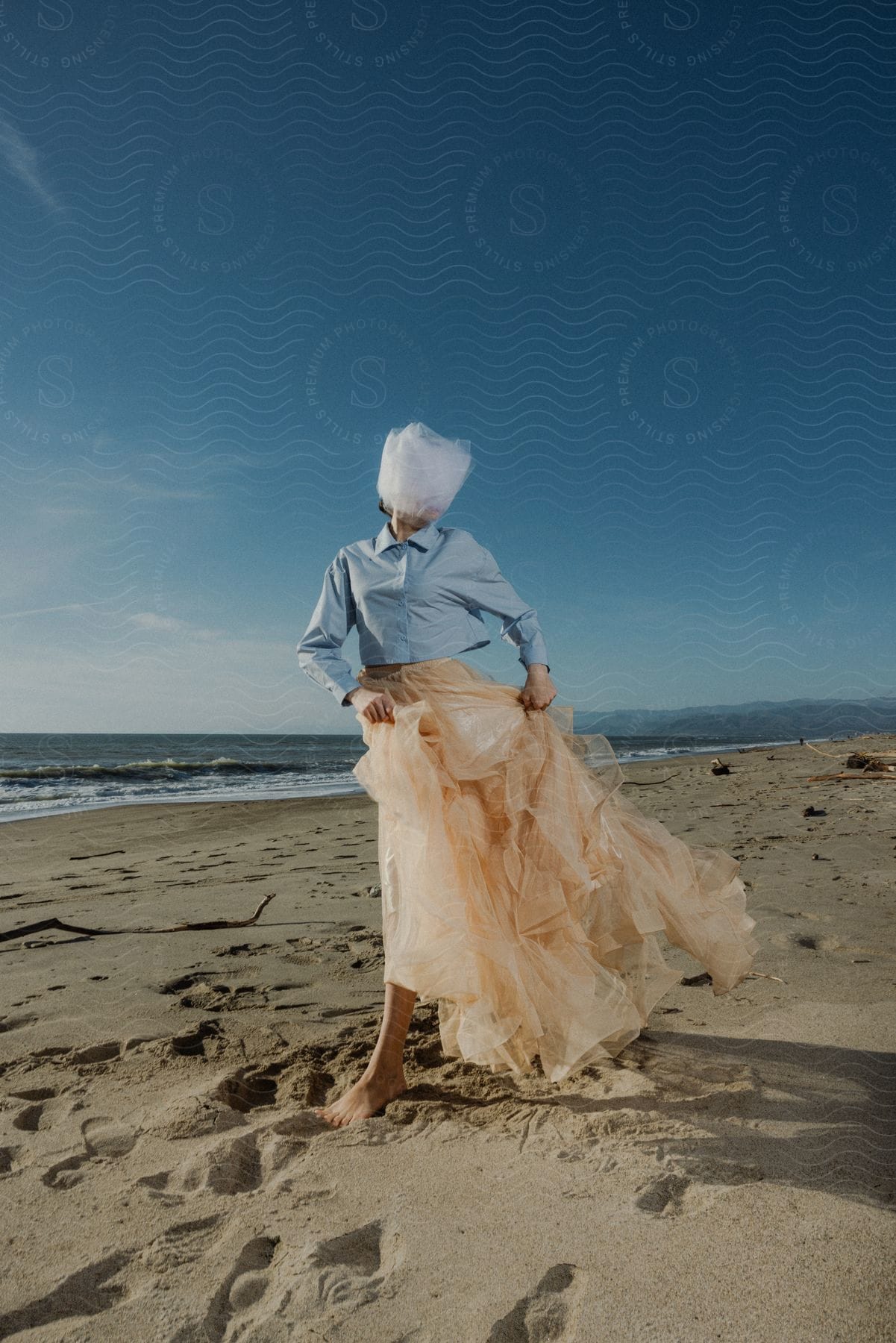 Woman with a covered face wearing a layered skirt stands on the beach barefoot.