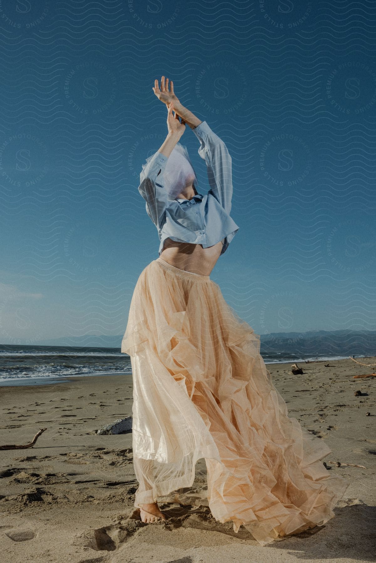 lady in a gown and light-blue crop top stands by the beach with both arms raised together at daytime