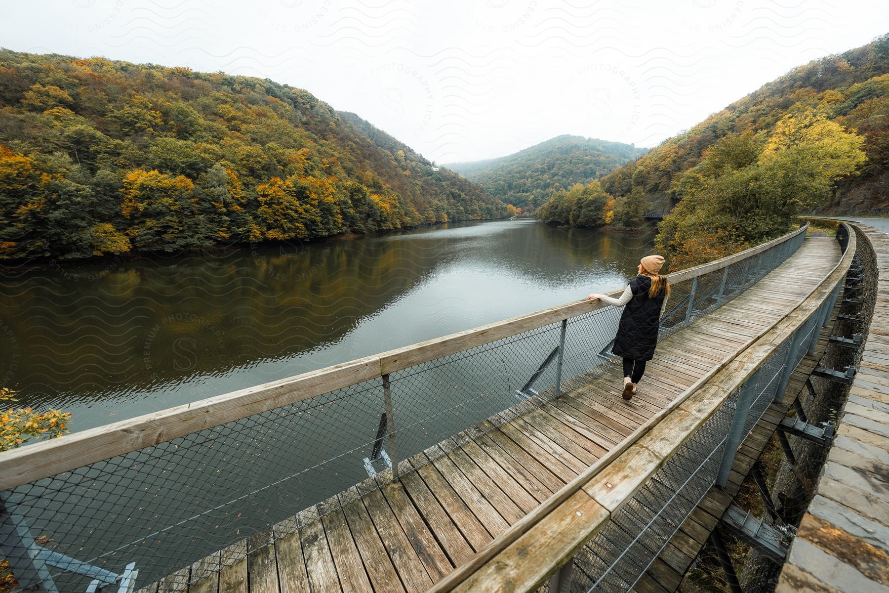 A woman, dressed in a brown hat and a long black vest, strolls across a bridge spanning a tranquil lake.