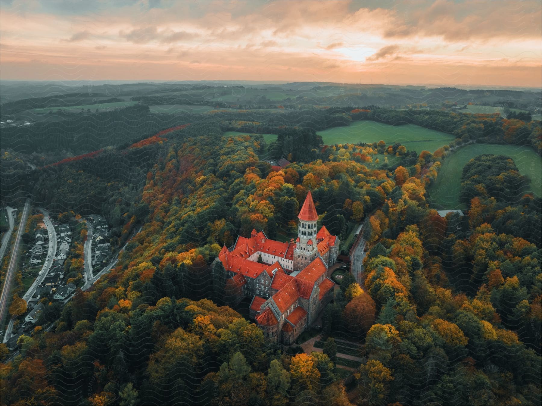 A red-roofed church sits on a hillside overlooking a small town, with trees turning yellow in the foreground and a sunset in the distance.