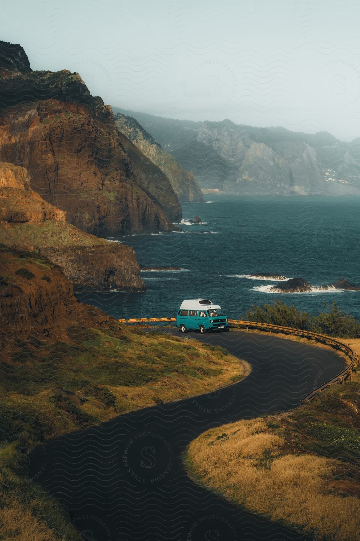 Van drives along coastal highway winding between mountain cliff and the ocean.