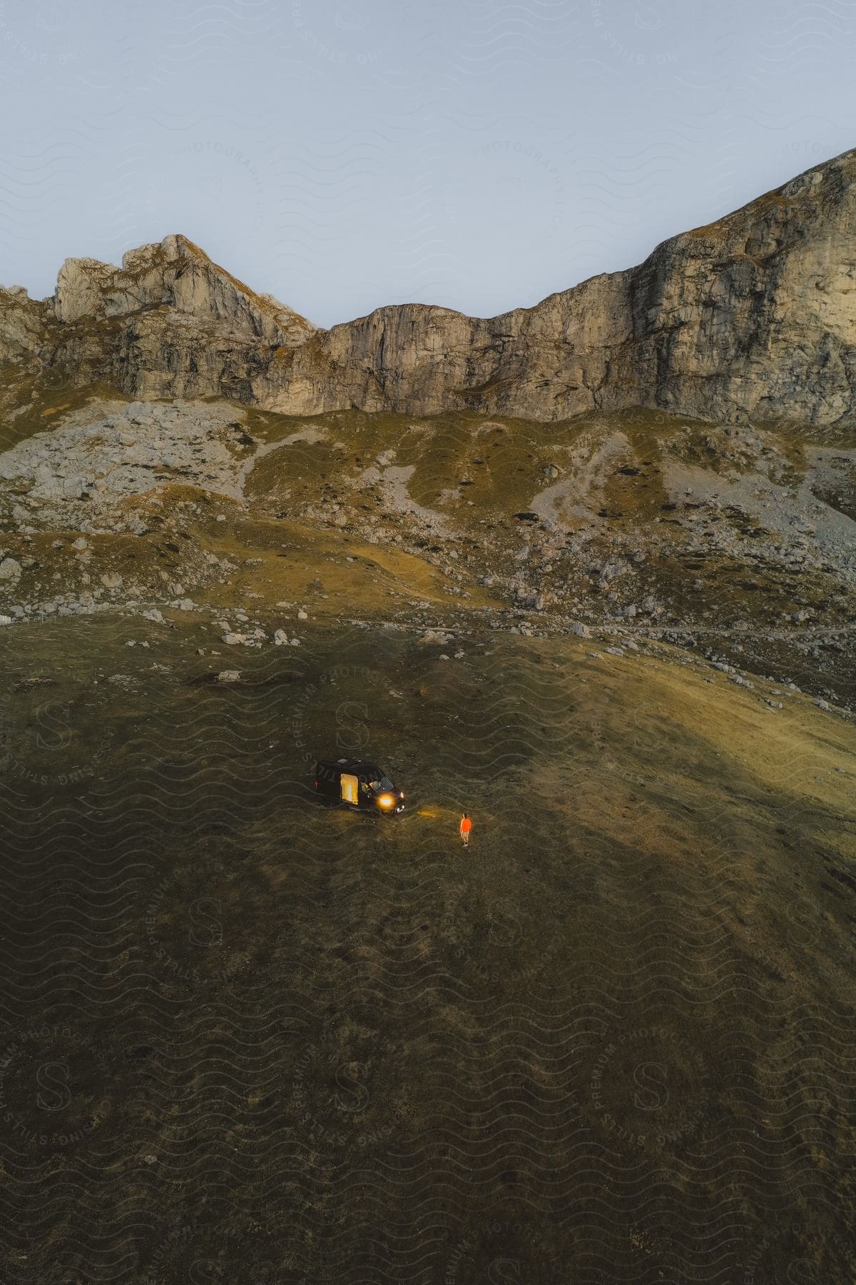 Person camping near mountains stands in front of headlights of van.