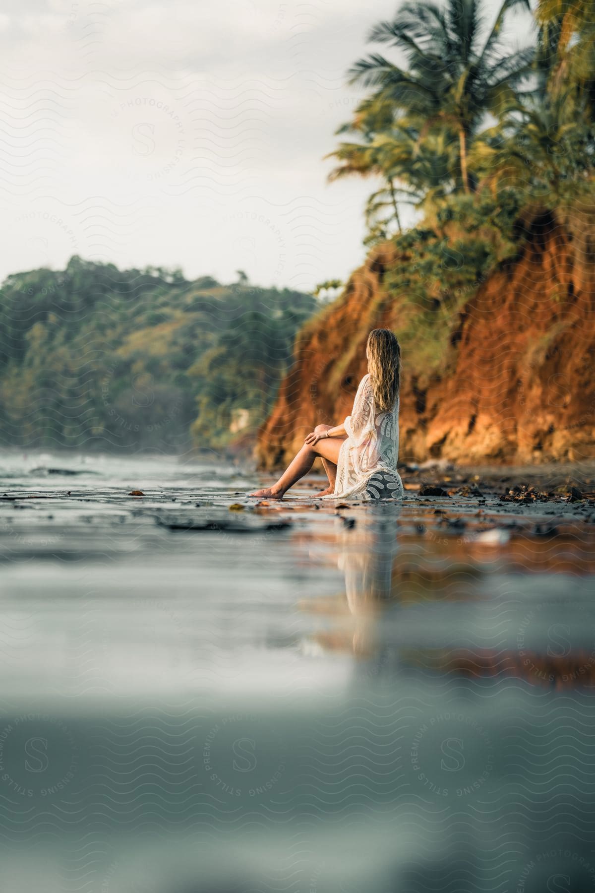 A woman sits on a rock in the water looking at the coastal promontory