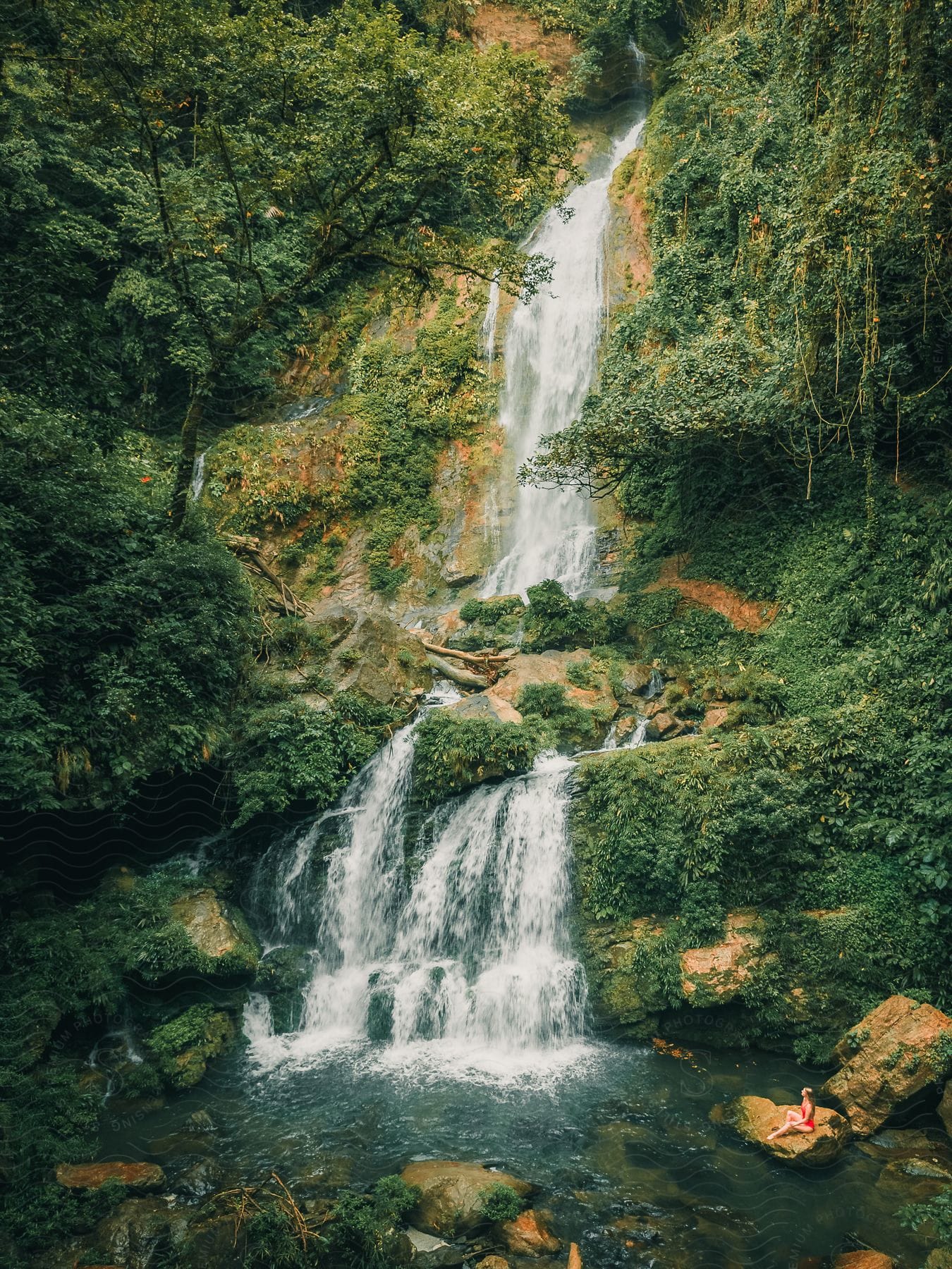 Woman sitting on a rock at the base of a multi-tiered waterfall surrounded by greenery.