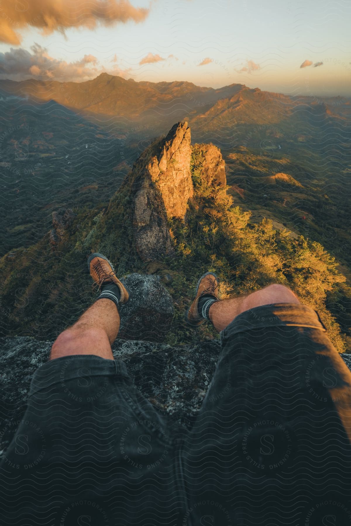 Person's perspective sitting on a mountain edge overlooking a rugged peak and valley during sunset.
