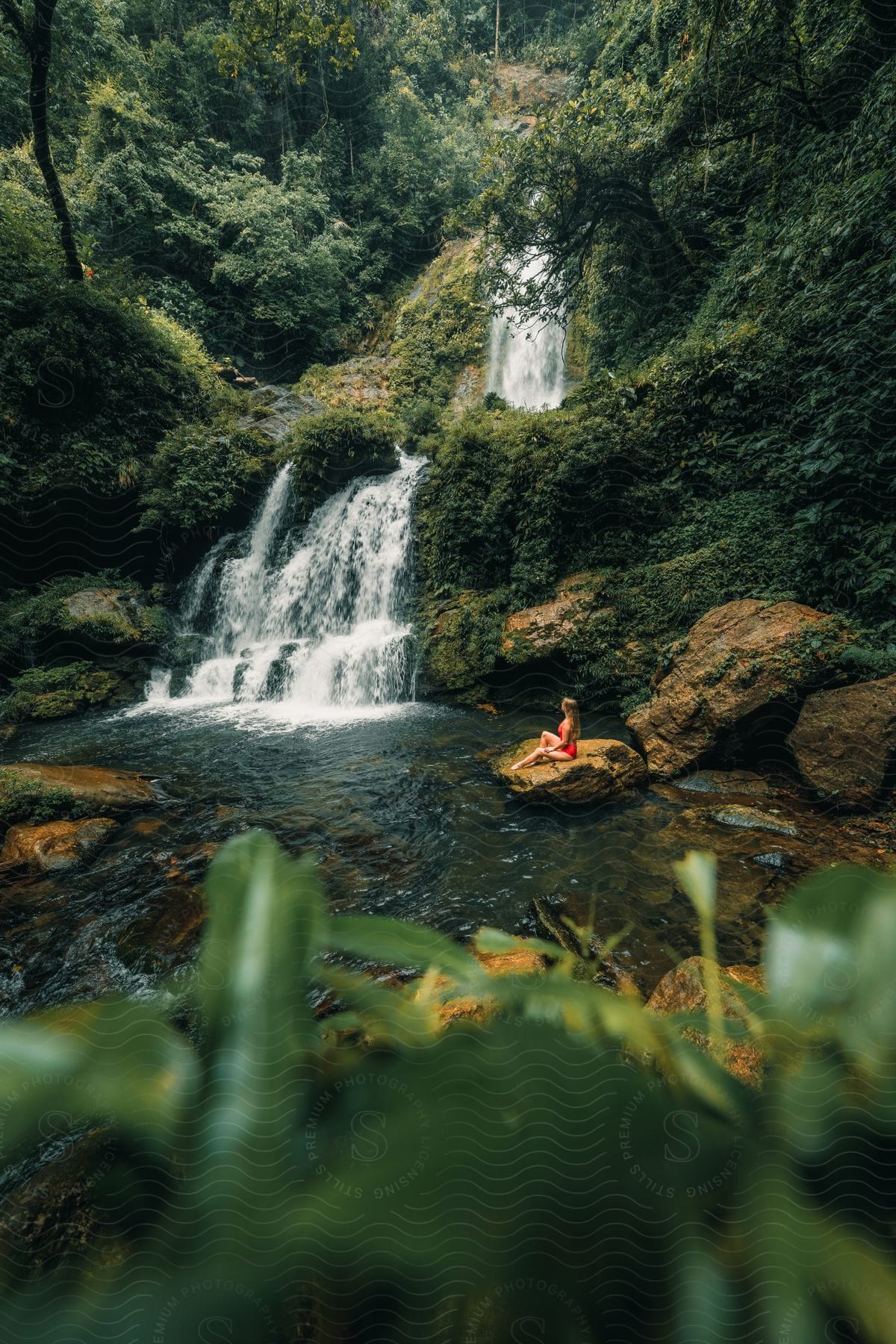 A Woman In A Red Bathing Suit Sits On A Rock In The Water Watching The Waterfall