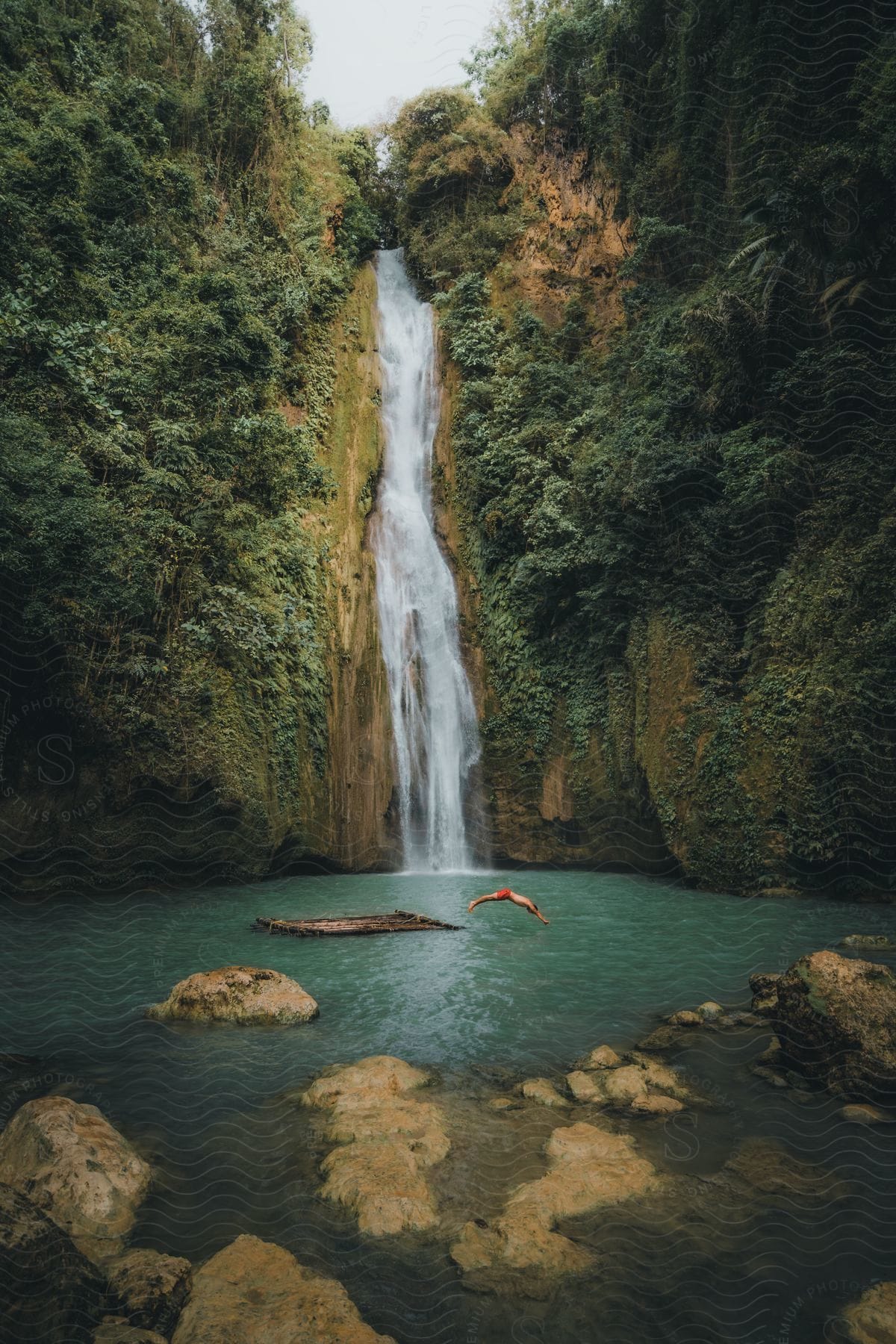 Natural landscape of a waterfall on a mountain with a man jumping into the greenish water.