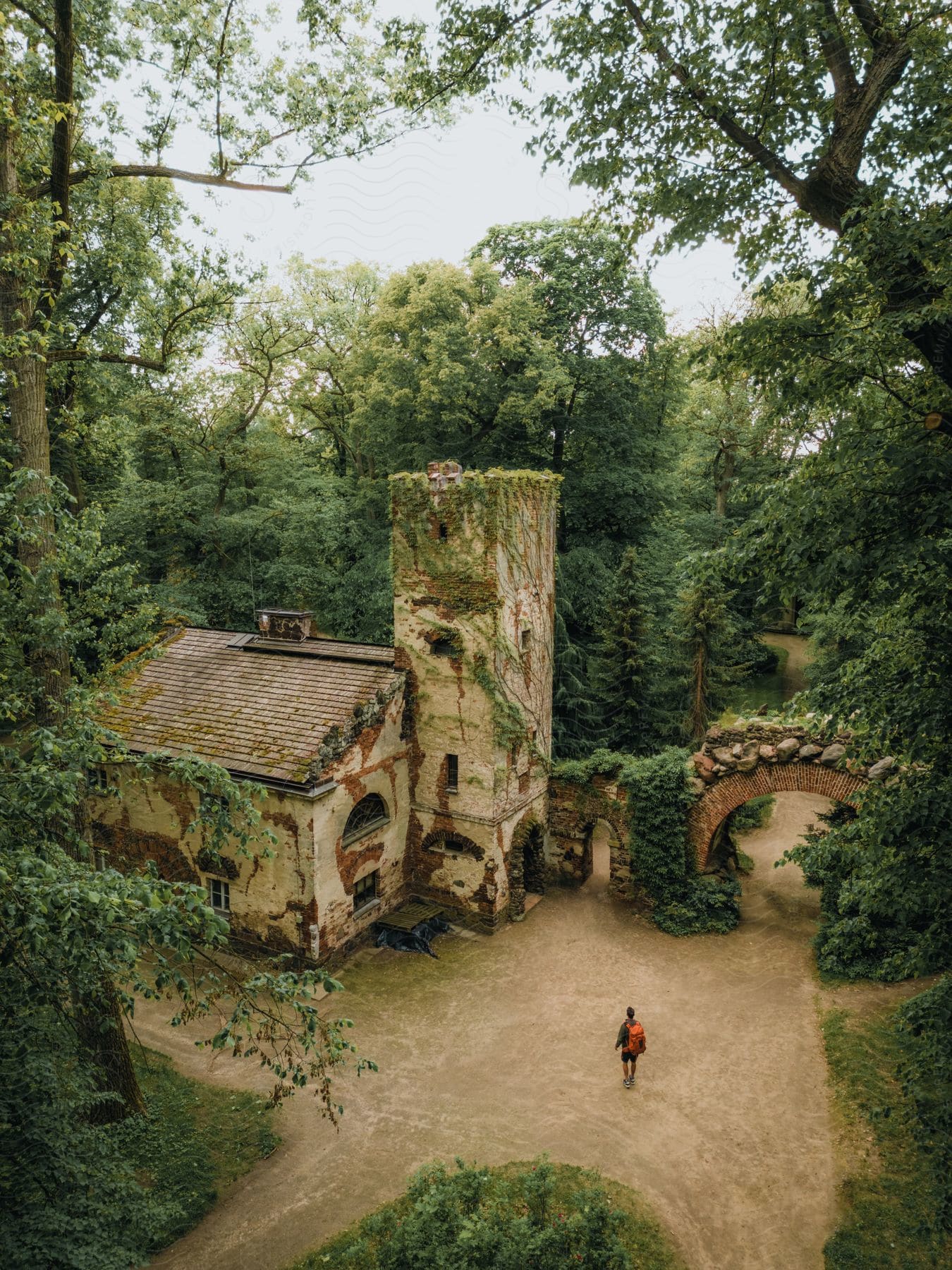 An ancient and partially ruined building is surrounded by lush greenery, with parts of its walls covered in ivy. A person walks near the building.