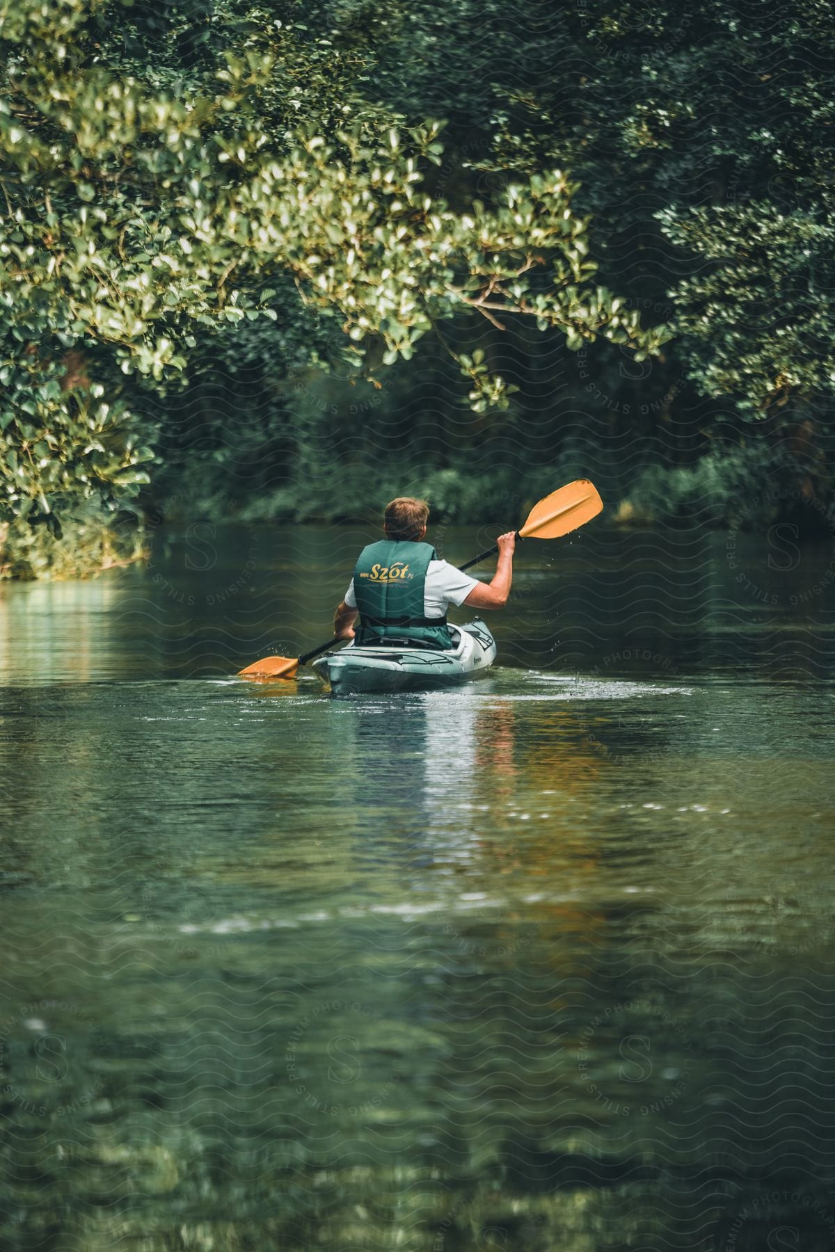 A man is backwards in a kayak wearing a green vest and orange paddle while sailing on a river