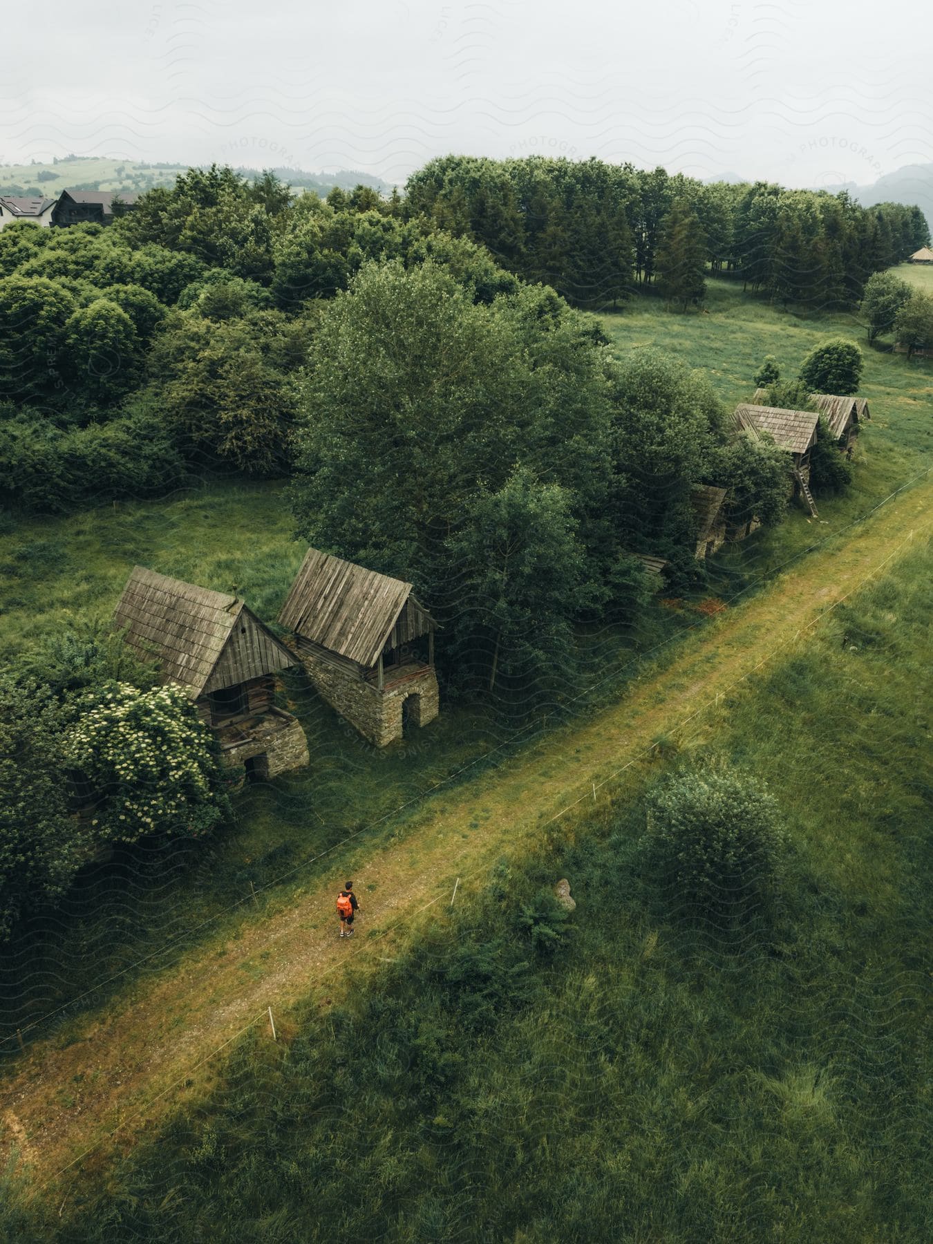 A person in red waling a path flanked by rural wooden houses in the countryside.