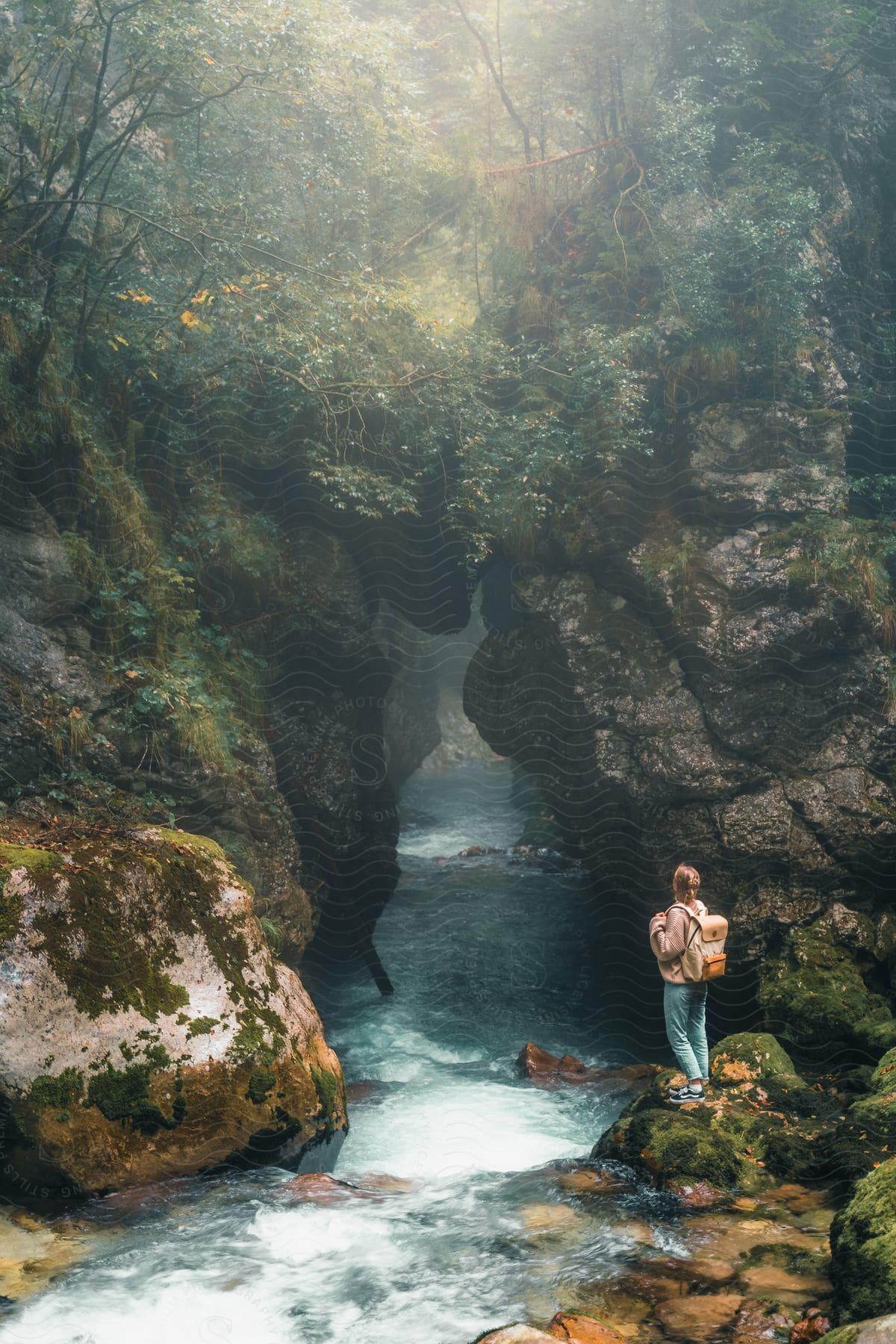 A hiker with a backpack stands on a rock near a cliff overlooking a flowing stream