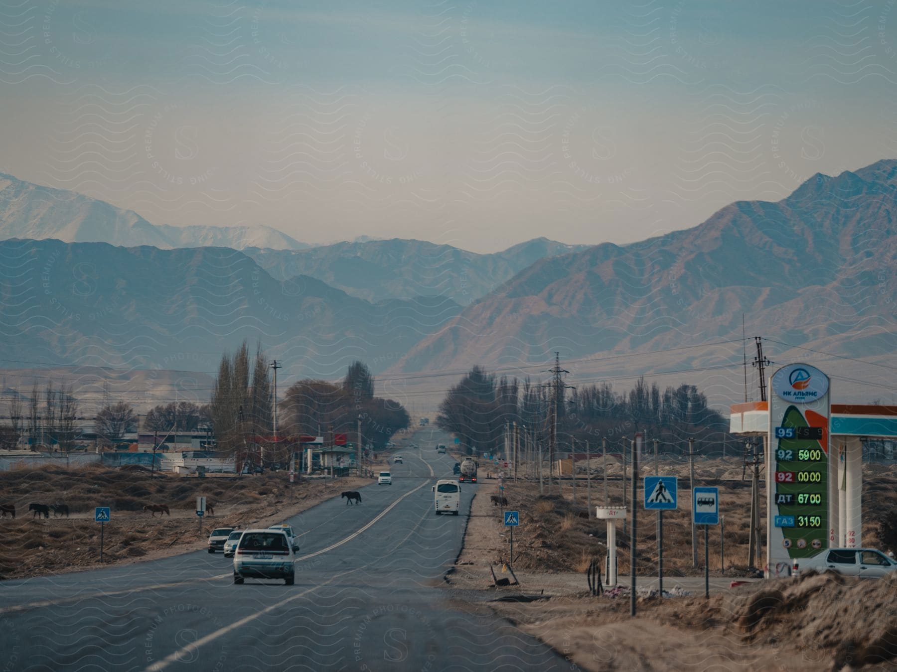 Vehicles travel on a highway with distant mountain views, utility poles, and grazing horses along the roadside, under a hazy sky. A fuel station sign displays prices in the foreground.