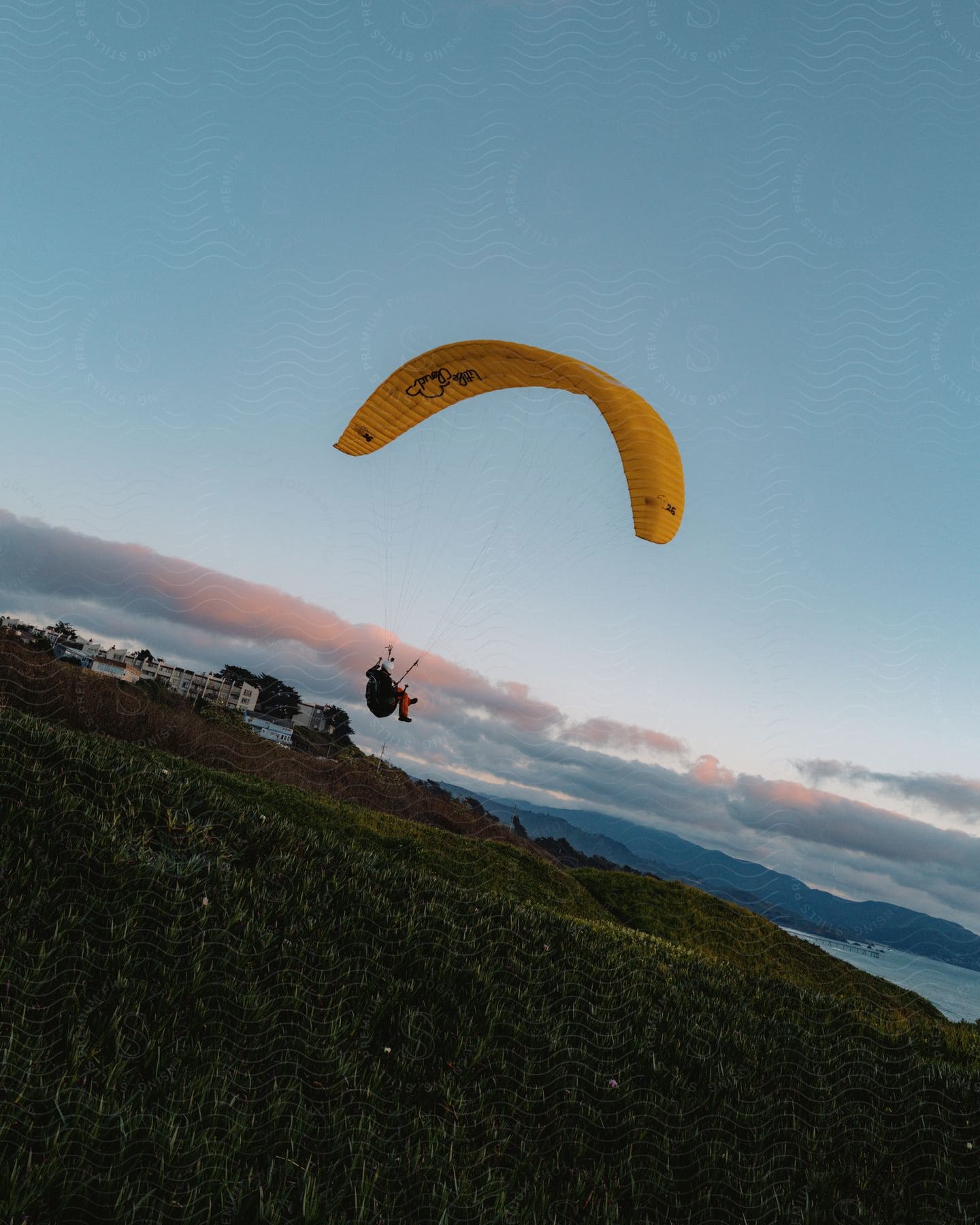 A yellow parachute carries someone in orange pants and a white helmet paragliding down a hill at sunrise or sunset.