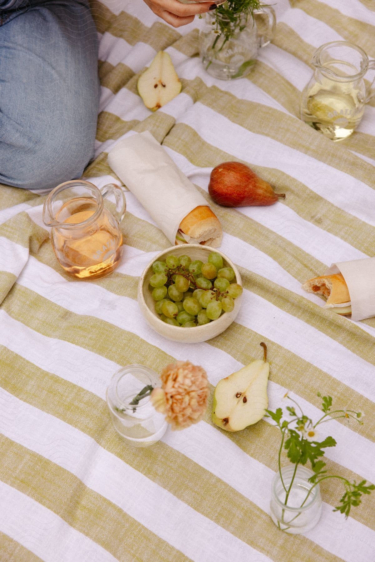 A person sitting on a picnic blanket with drinks, fruits, and sandwiches