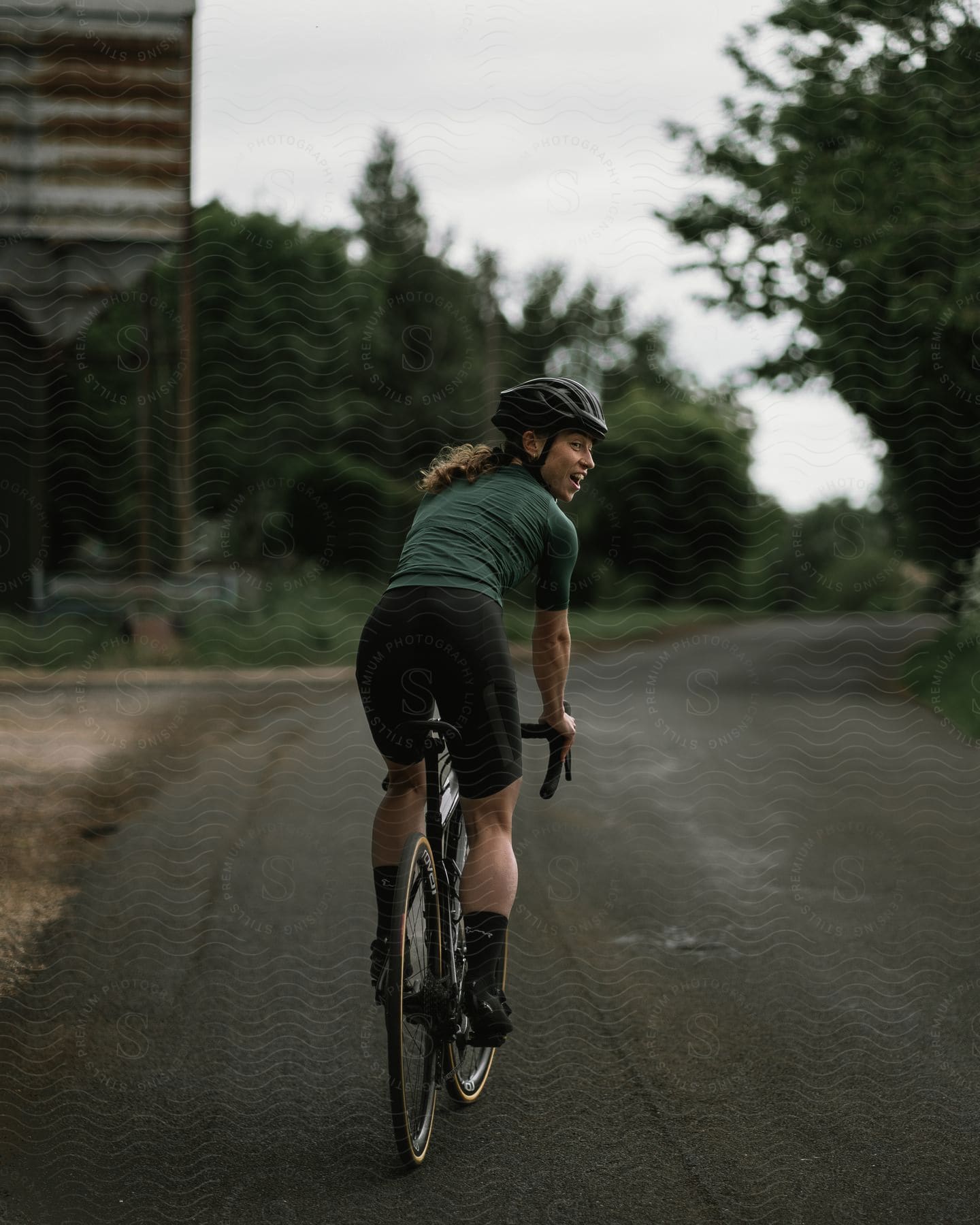 A cyclist wearing a helmet, a green shirt, and black shorts looks over her shoulder while riding a bicycle