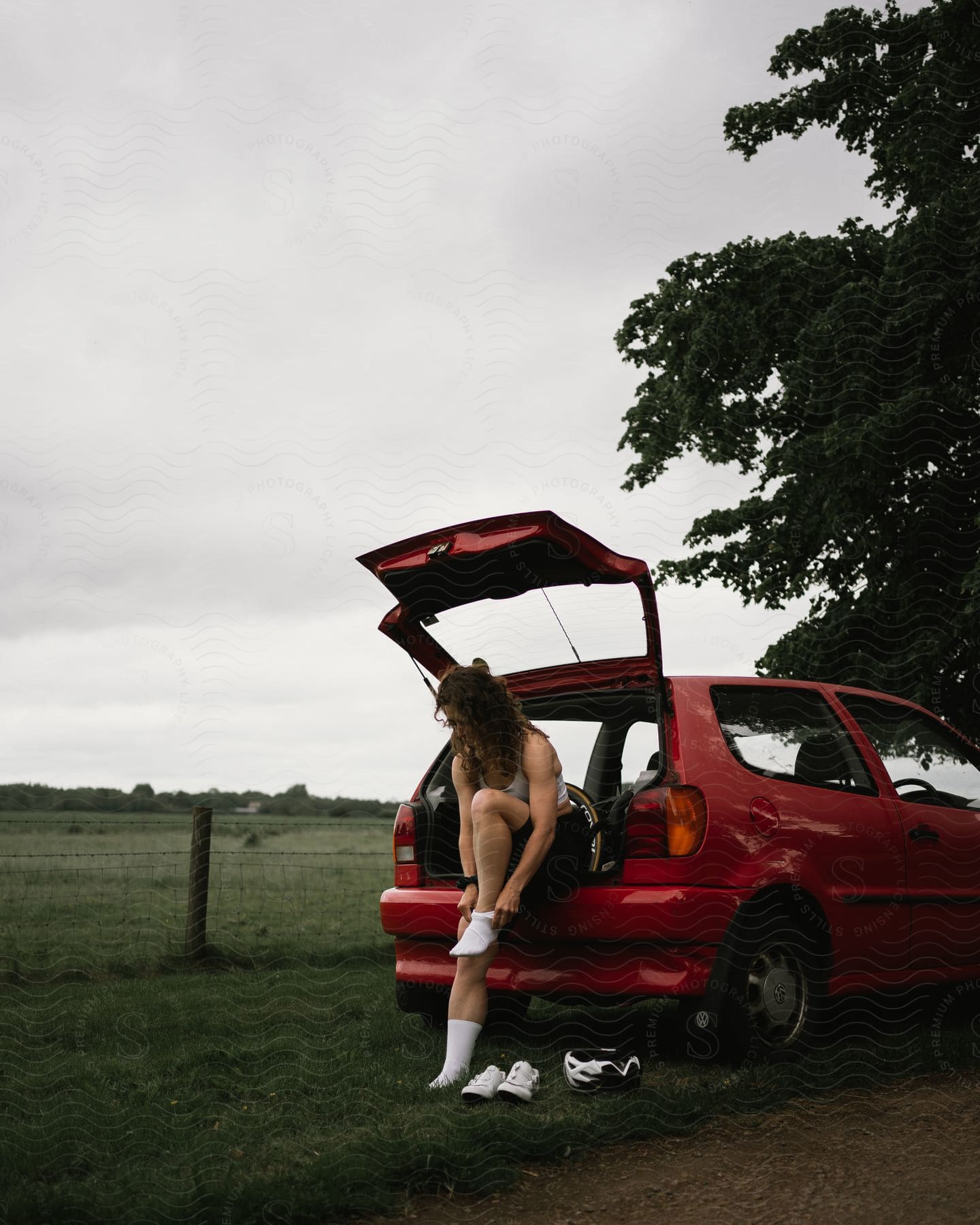 A woman sits in the trunk of a red car, tying their shoe, with sports gear on the ground beside them.