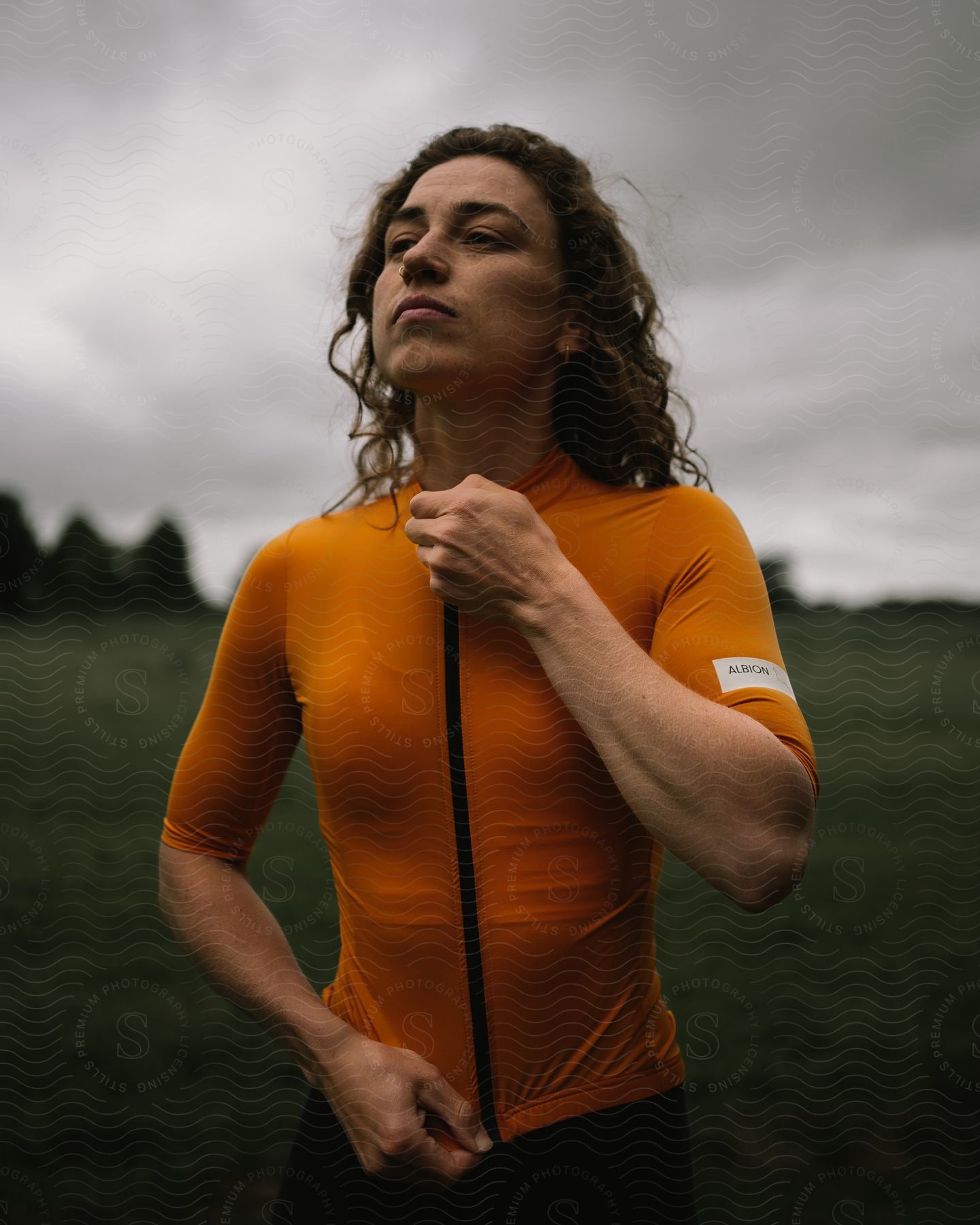 Model zipping up orange top and cycling jacket while standing in a field on a cloudy day