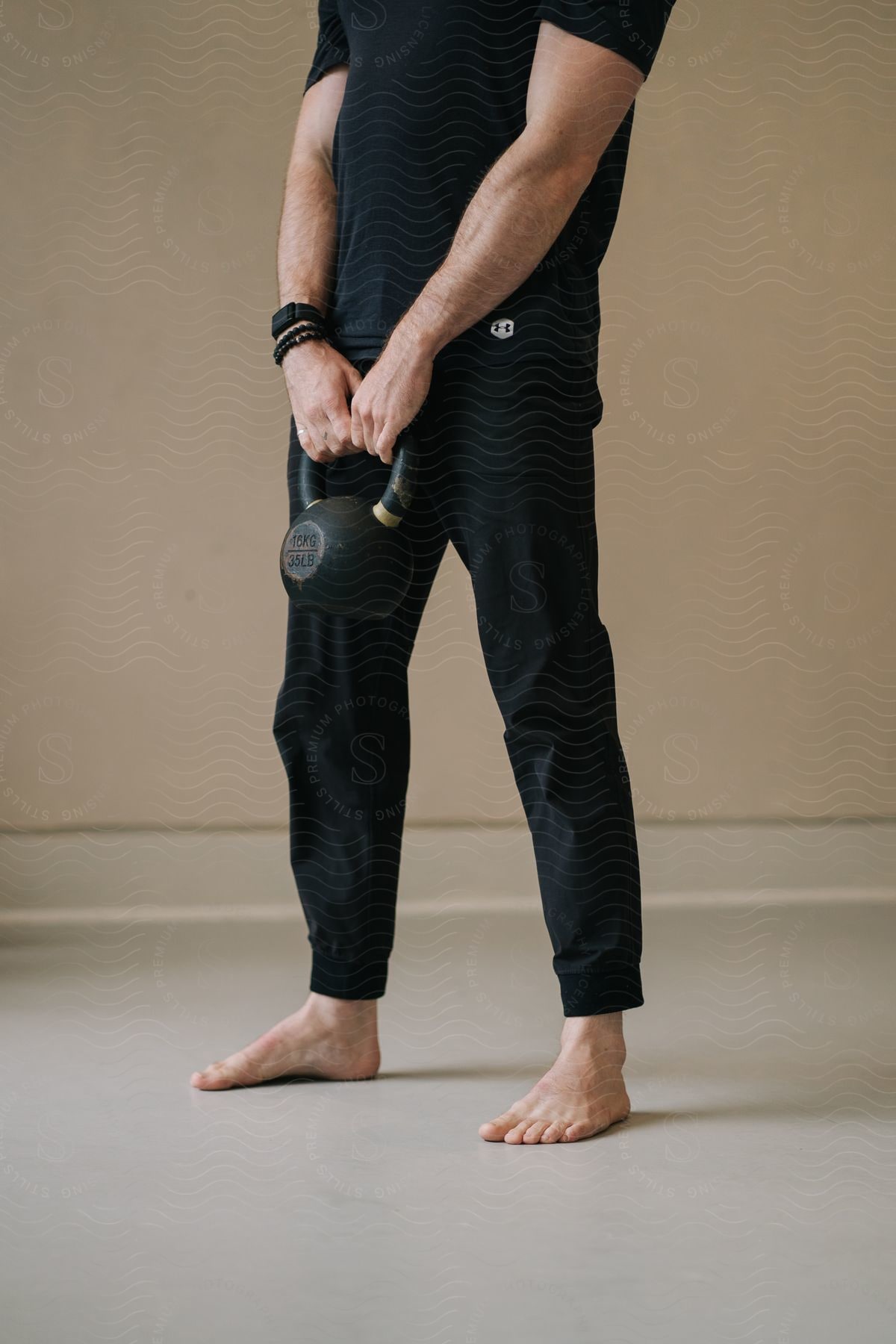 Man holding a kettlebell, wearing a black t-shirt and athletic pants, standing barefoot