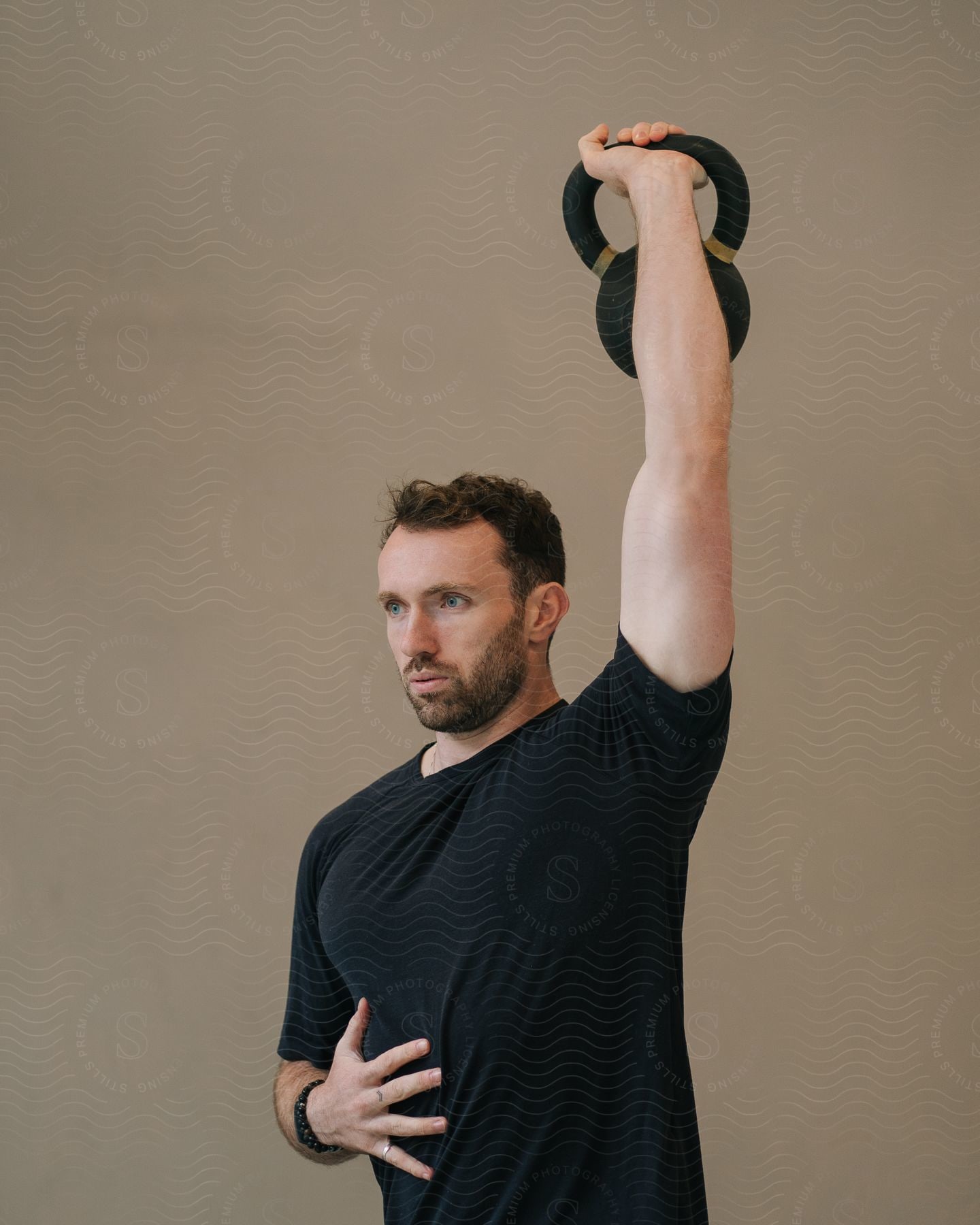 A bearded man wearing a black shirt is lifting a gym weight upwards with his left arm.