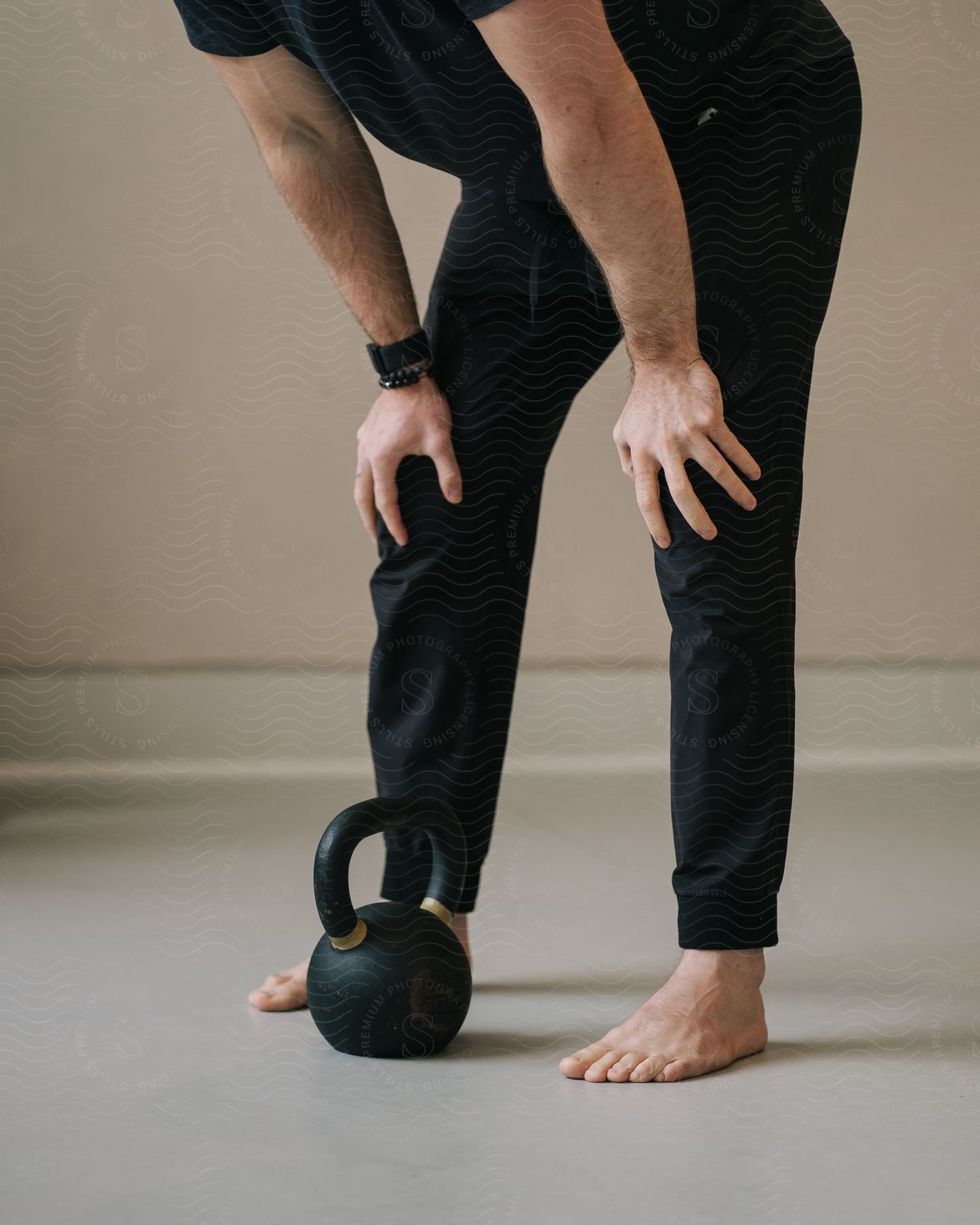 Lower body of a man in black athletic wear with hands on knee, standing by a black kettlebell on the ground.