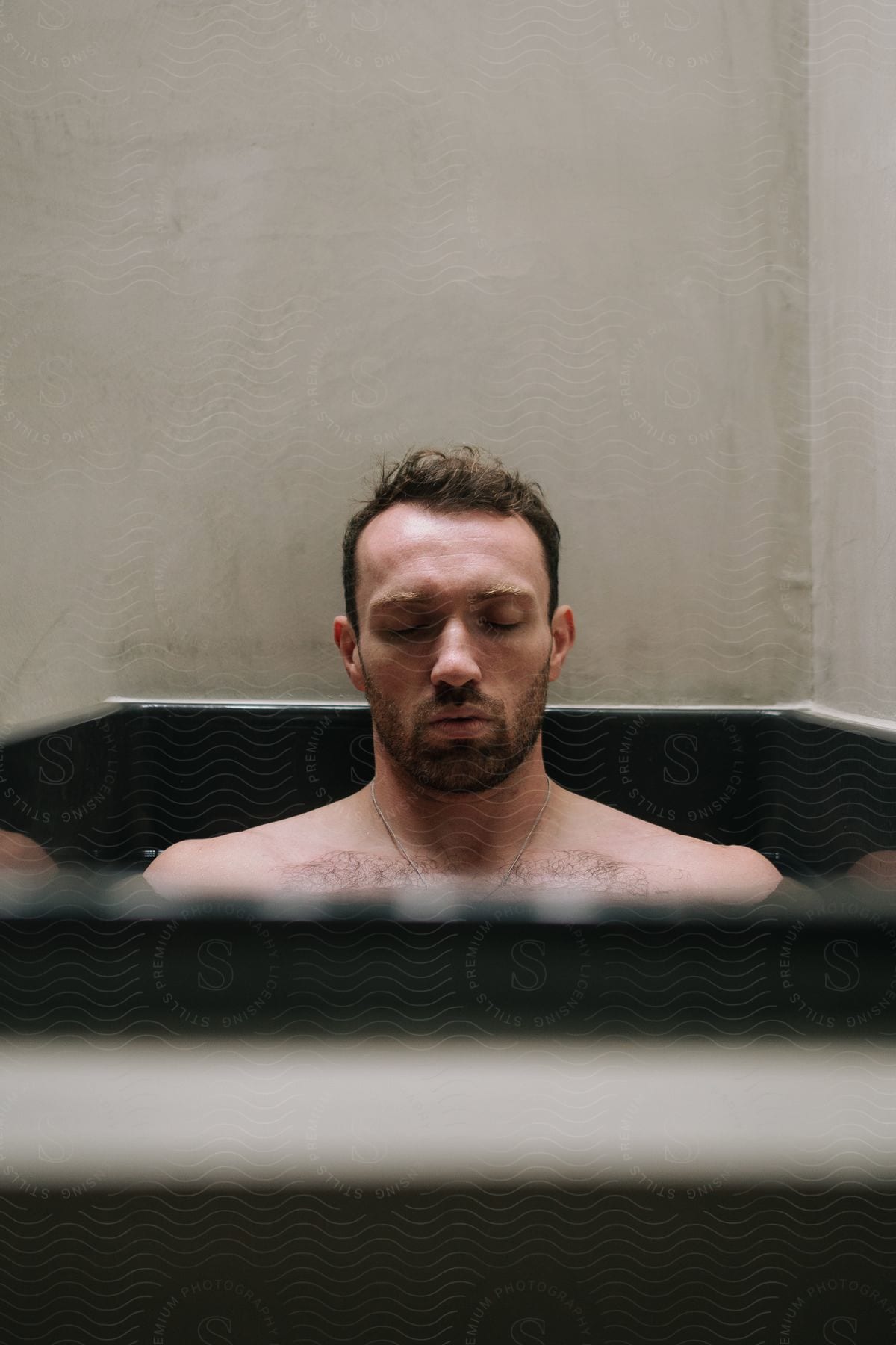 Man closes his eyes while he sits in an ice bath in a black tub.