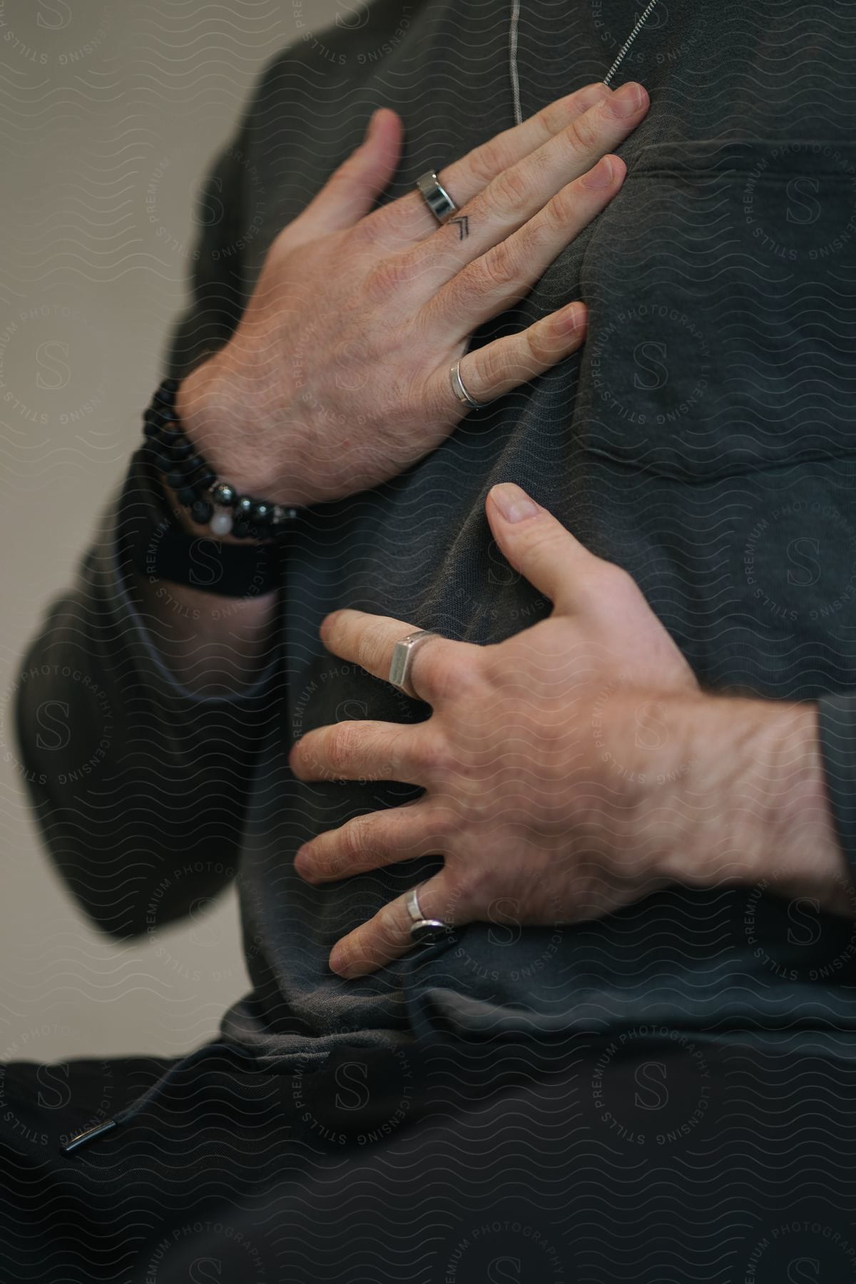 A person's hands holding their chest, with several rings and a black bracelet with large beads.