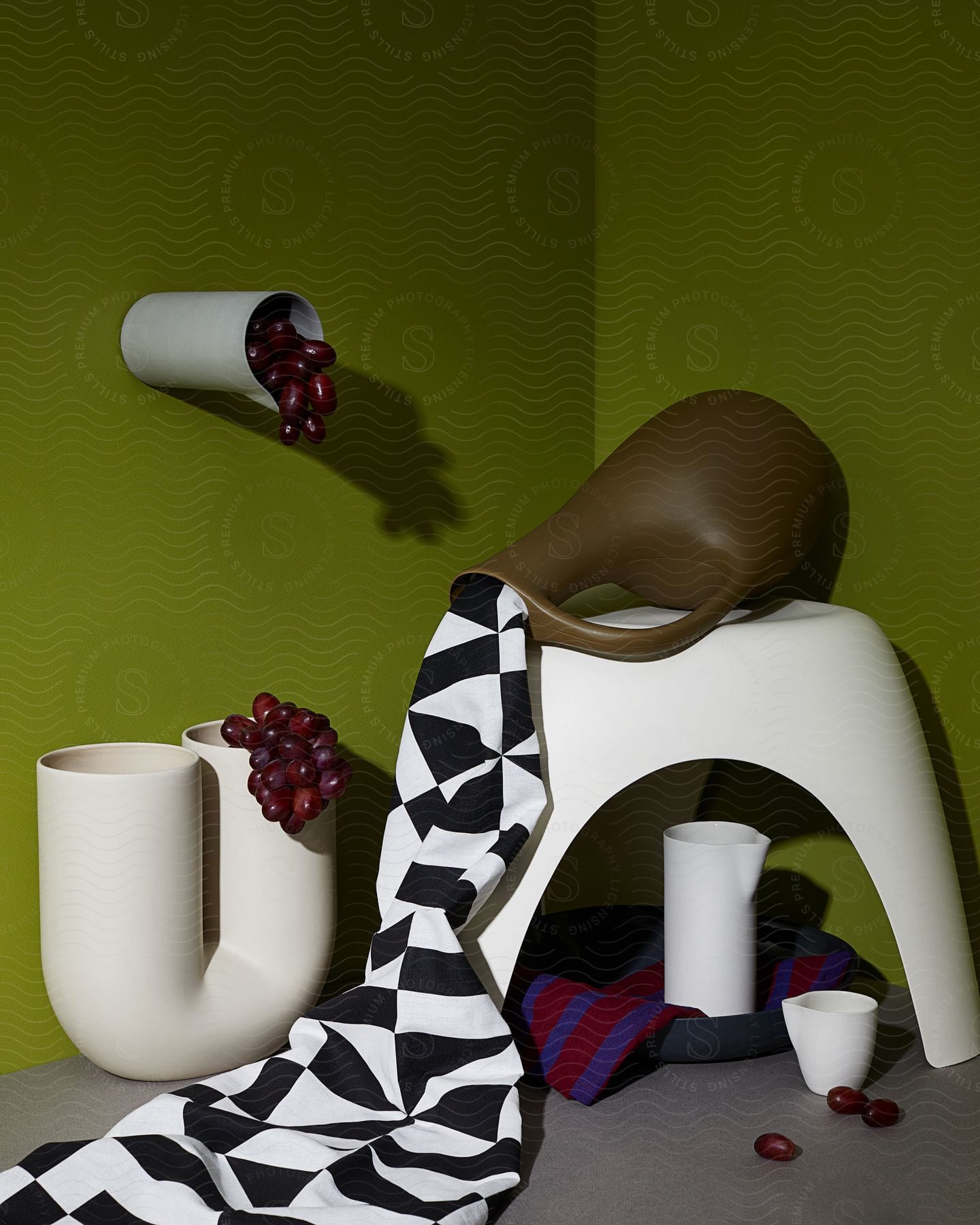 A white pipe with grapes coming out of its hole and there is a brown jug on a white table with a checkered fabric coming out of it.