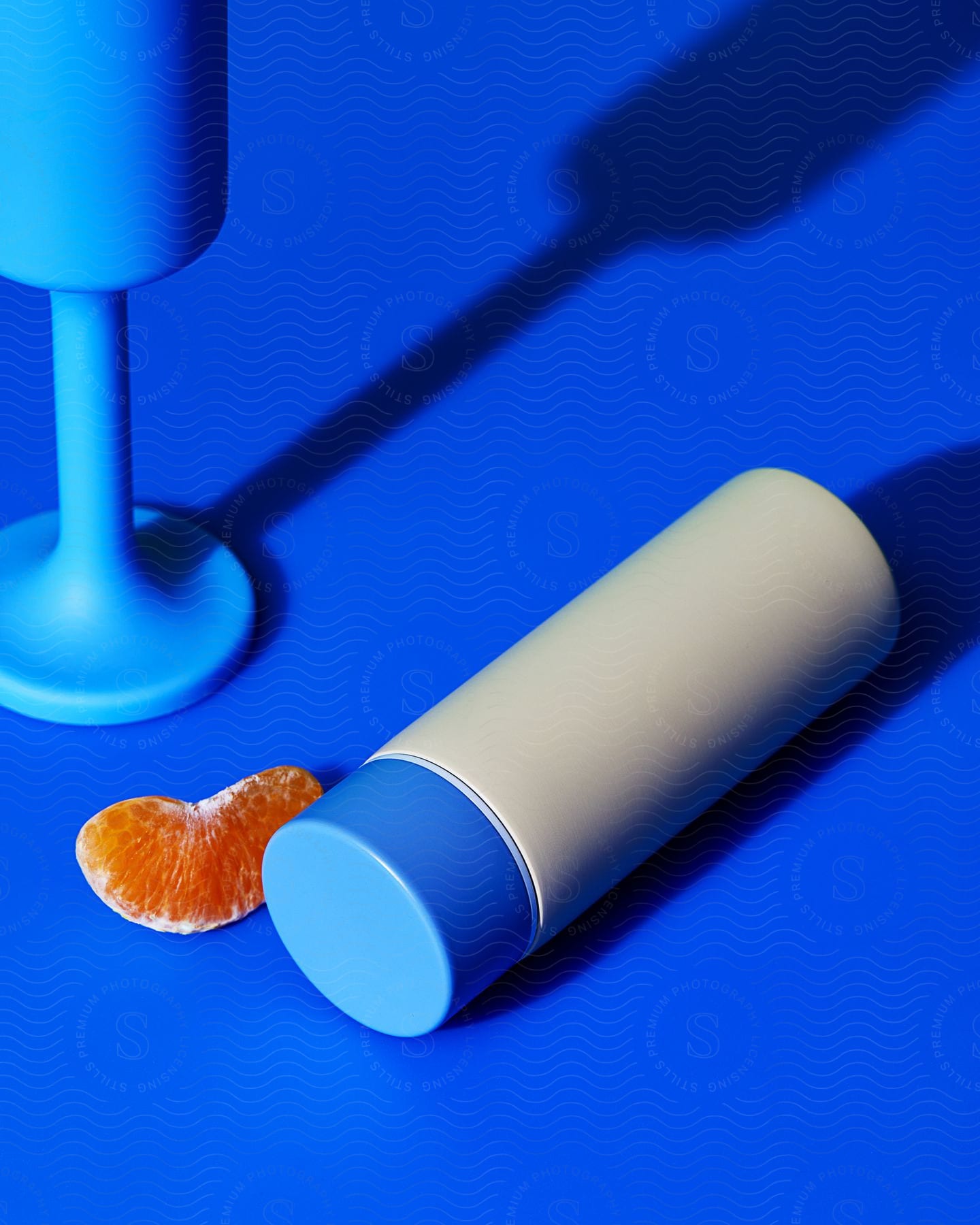 A white and blue cylindrical container lies on a vibrant blue surface and next to a tangerine slice there is a blue stemmed tall glass partially visible in the upper left corner.