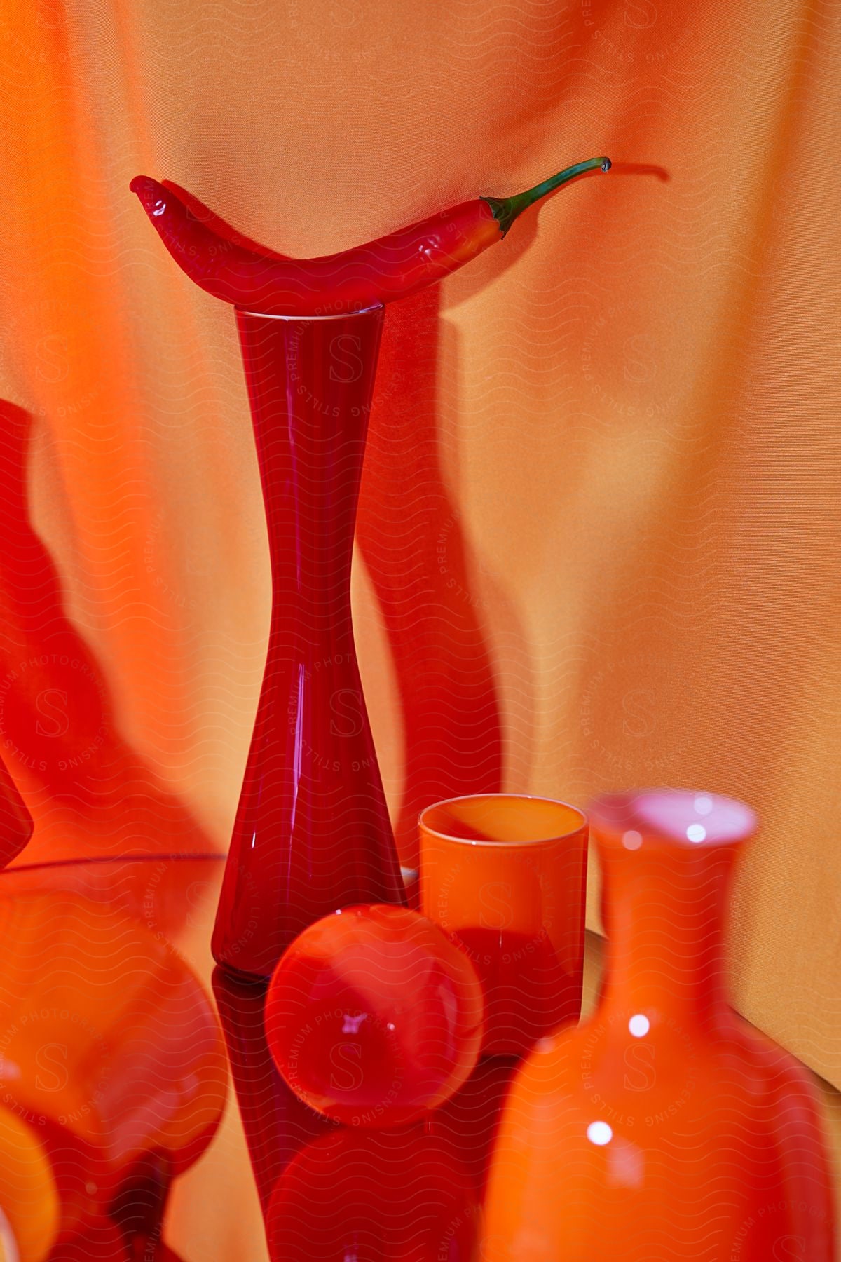 Collection of red and orange glassware on reflective surface with orange fabric background. A red chili pepper rests on top of a tall red vase.