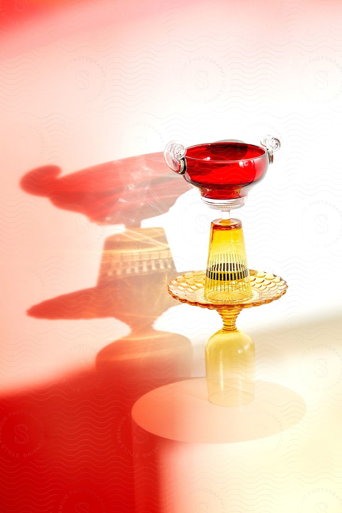 Red and clear glass compote on a yellow pedestal dish, against a gradient pink to white backdrop with shadows reflecting on surface.