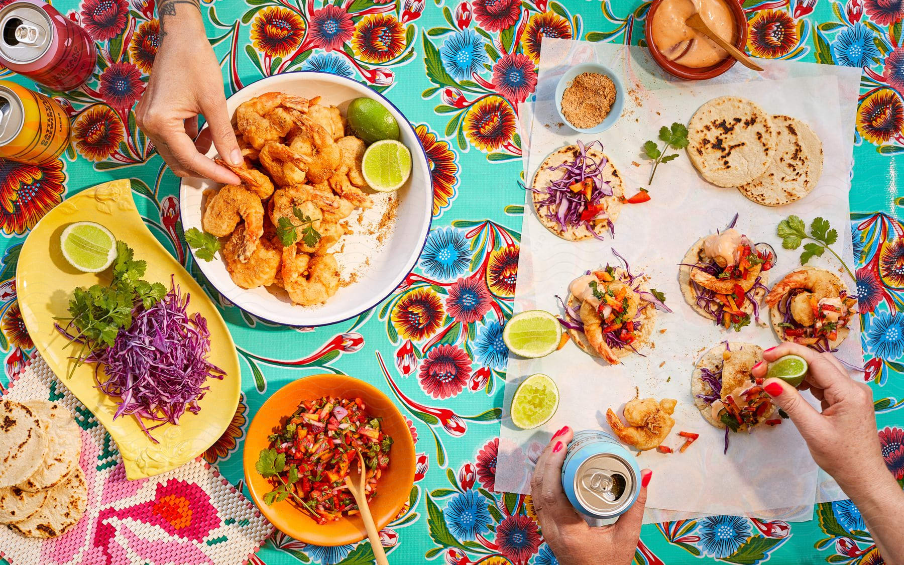 Colorful table spread with various Mexican dishes and hands serving and eating.