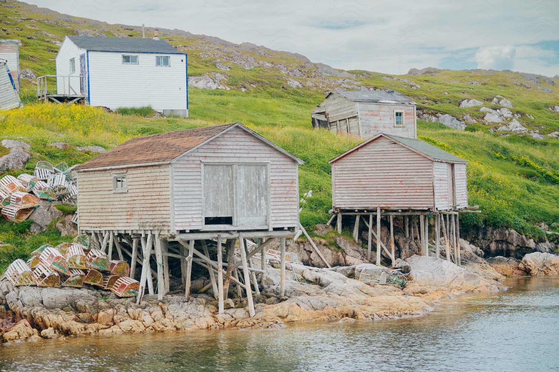 Fishing sheds on stilts along a rocky coastline with scattered lobster traps and a backdrop of green hills.