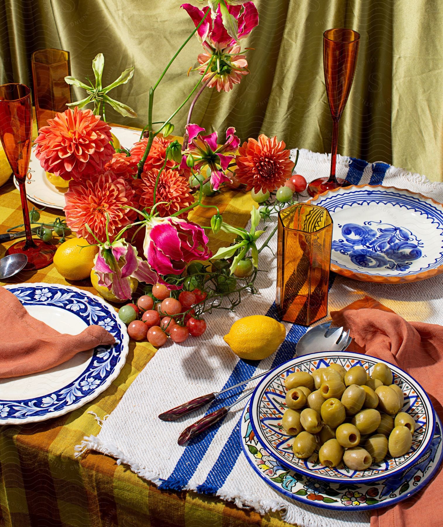 Olives in a bowl and meat on a plate, both on a table set for four with flowers and fruit as the centerpiece.