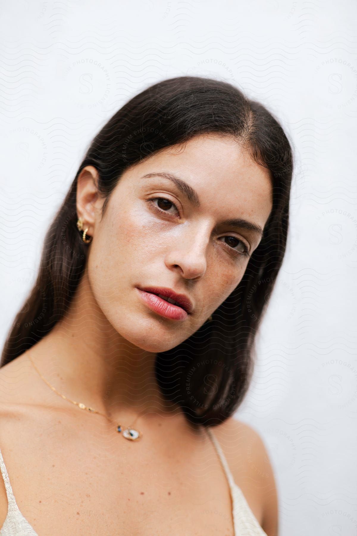 Close-up of a woman with dark hair and gold earrings, wearing a light-colored top and a gold necklace with a blue and white pendant, against a white background.