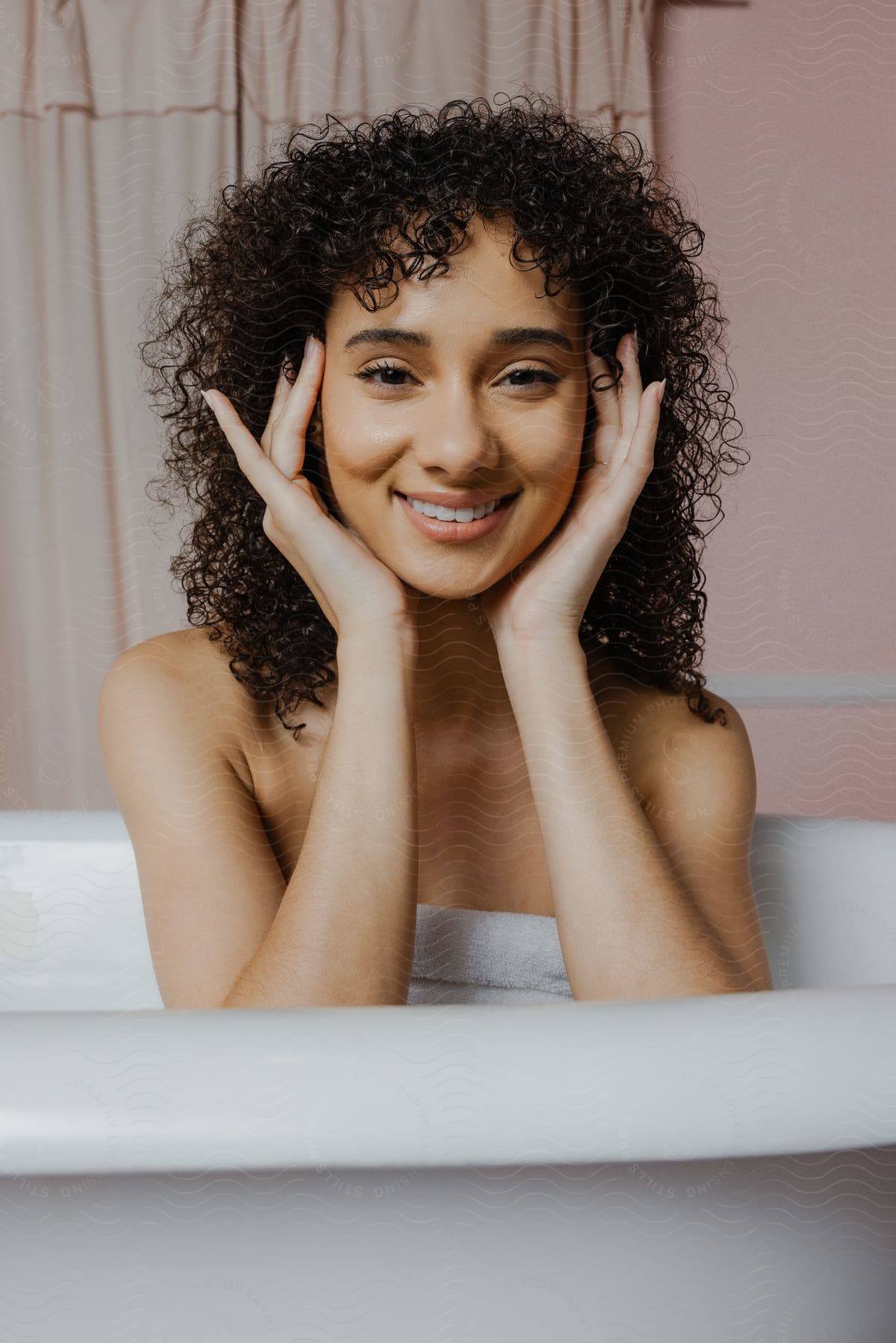 Woman in bathtub, hands on cheeks, smiling with curtains in background.