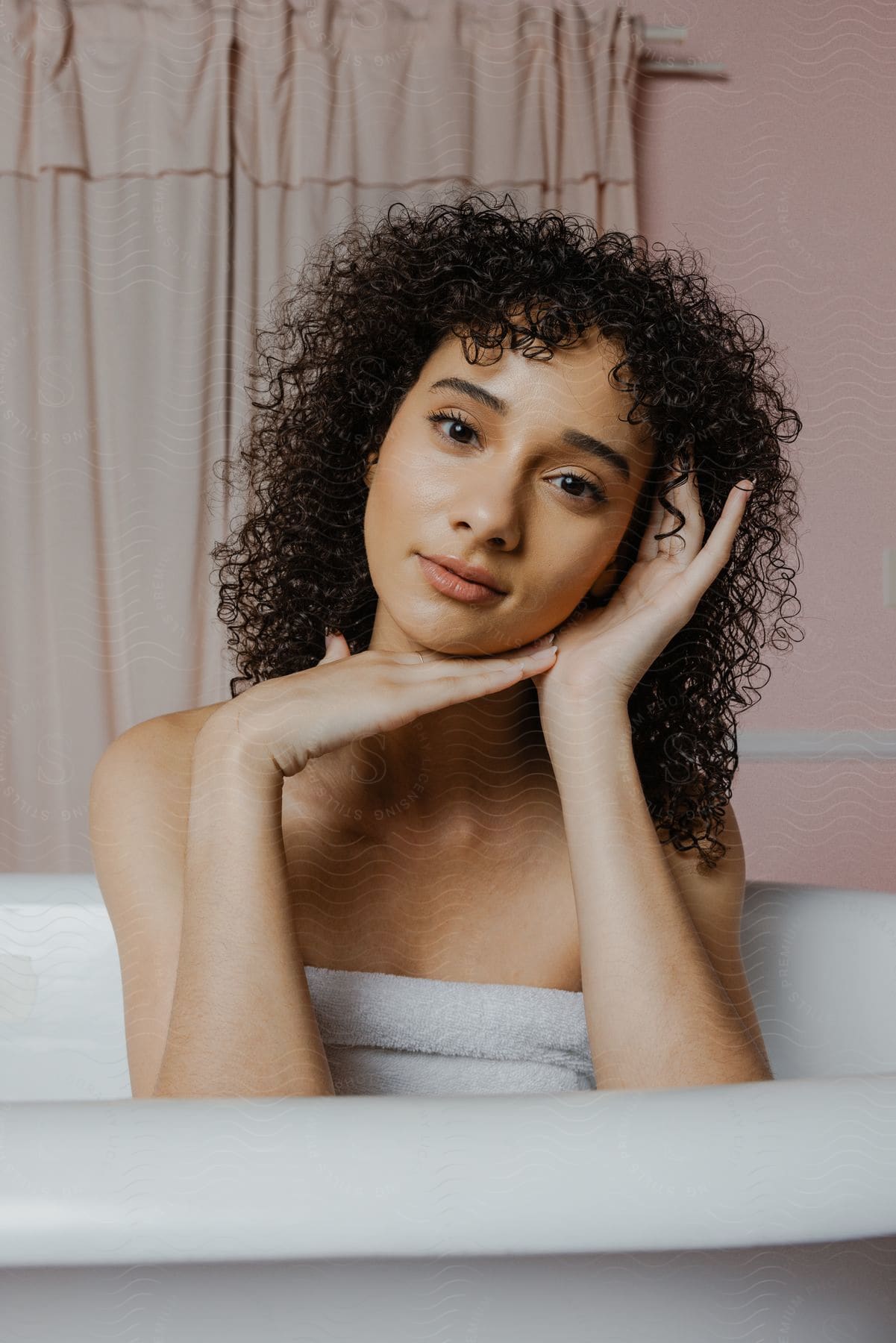 A woman holding her head with her hands is looking forward and is in the bathtub.