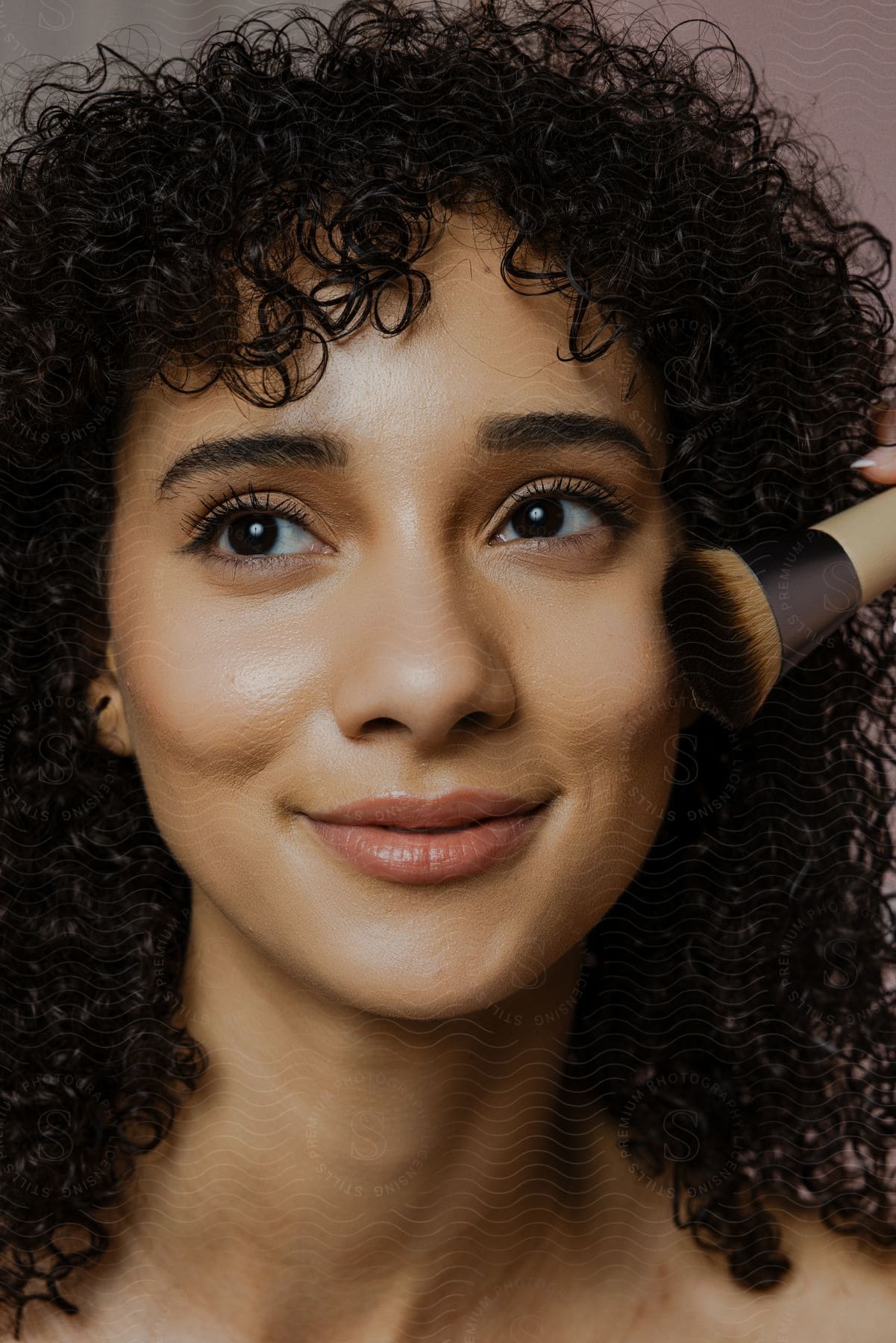 Portrait of a young woman with curly hair applying makeup brush to her face