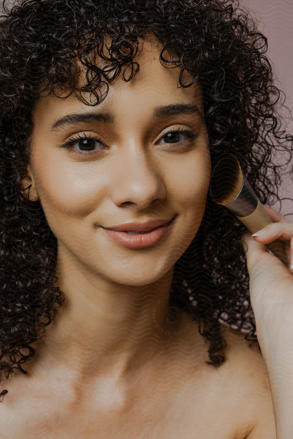 The face of a woman who is smiling with her lips and is holding a makeup brush close to her face and her hair is curly and dark.