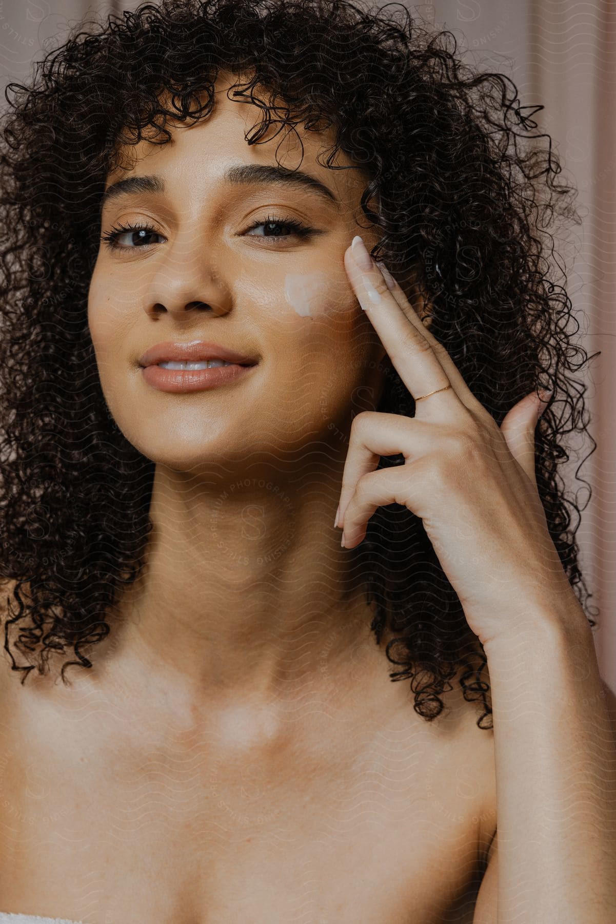 Woman with a slight smile and curly hair, applying cream to her face.