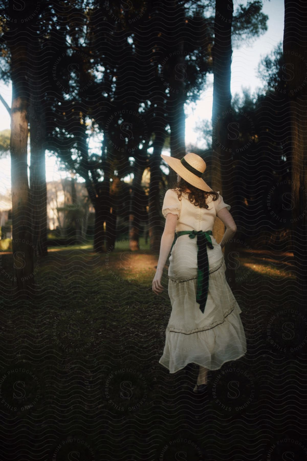 Stock photo of a person from behind, wearing a long white dress and a hat, in a park at dusk.