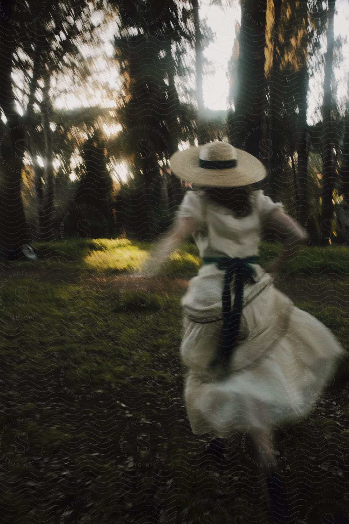 Woman with her back running through the garden surrounded by trees with a sun hat and a long white dress with a green ribbon at her waist.