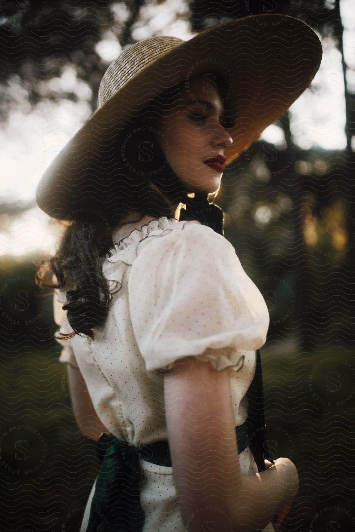 Woman in a white dress with green polka dots and sash wears a straw hat in the forest.