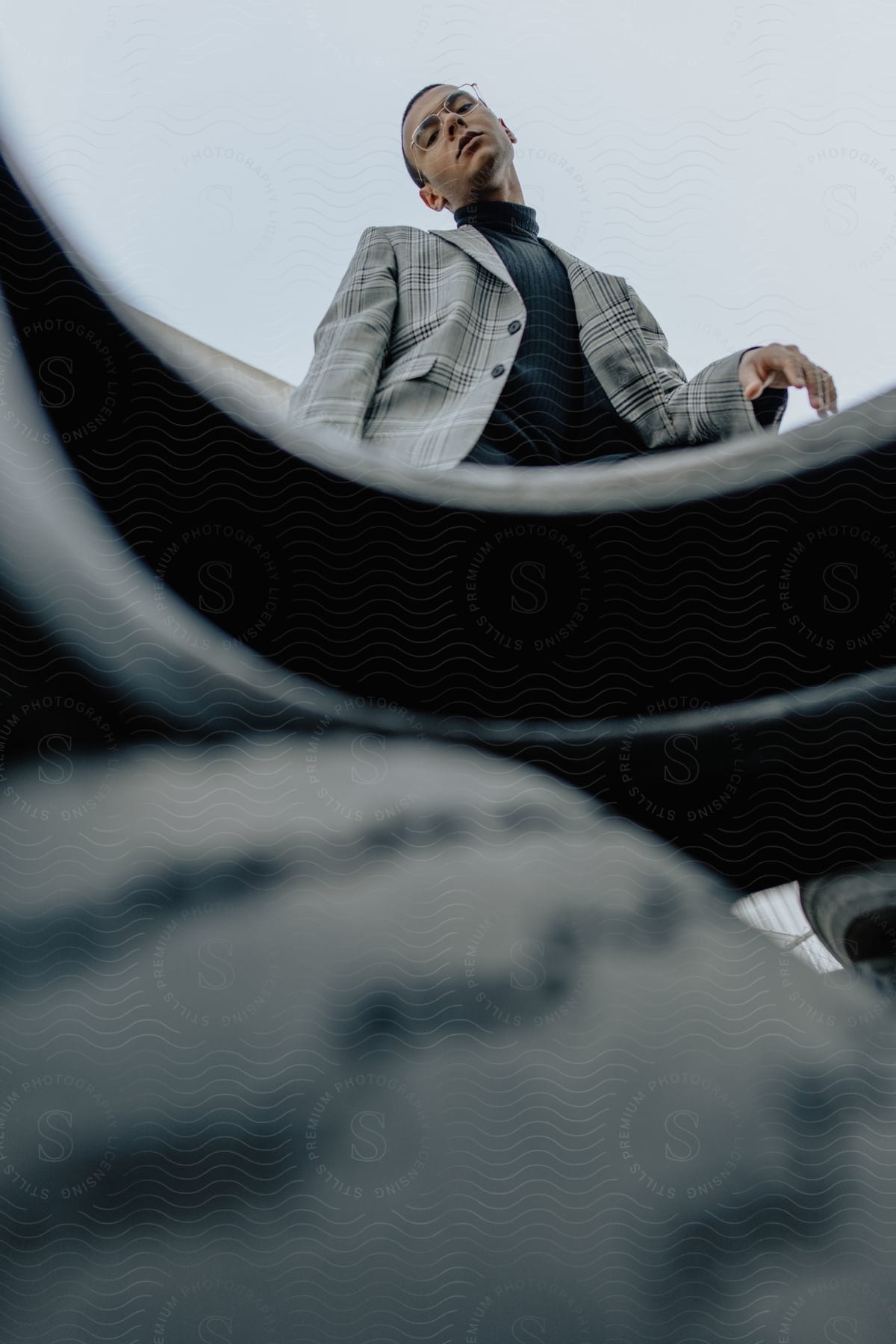 A man in glasses and a plaid jacket stands with his hand raised, looking straight ahead at a blurred tire below him.