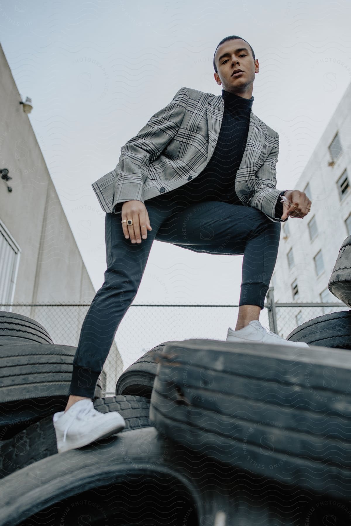 Man wearing a gray blazer jacket black shirt pants and white sneakers poses for the camera on tires