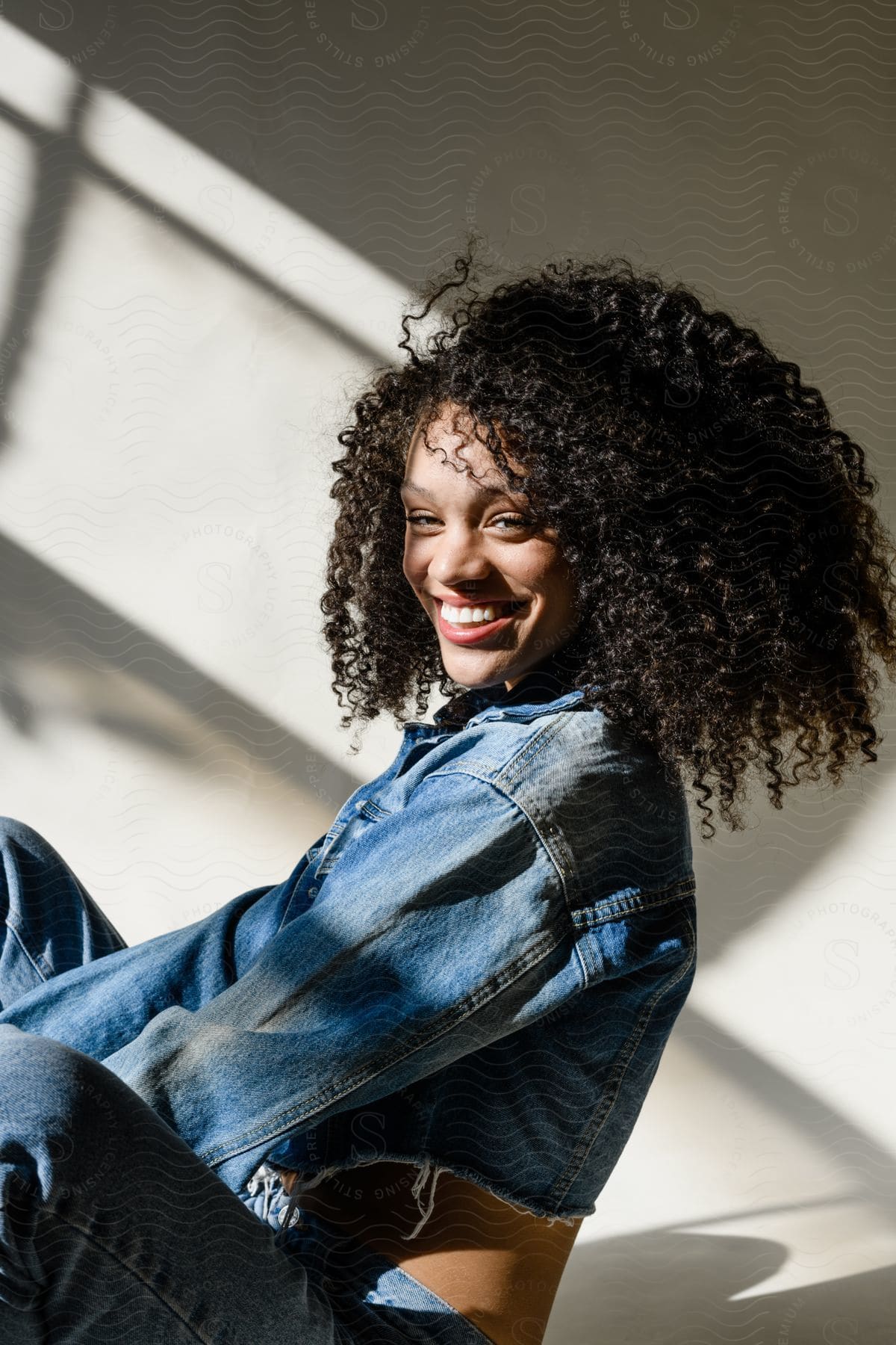 A woman with curly hair is smiling in a room with a concrete wall and is wearing jeans while the sunlight casts shadows on the wall in the background