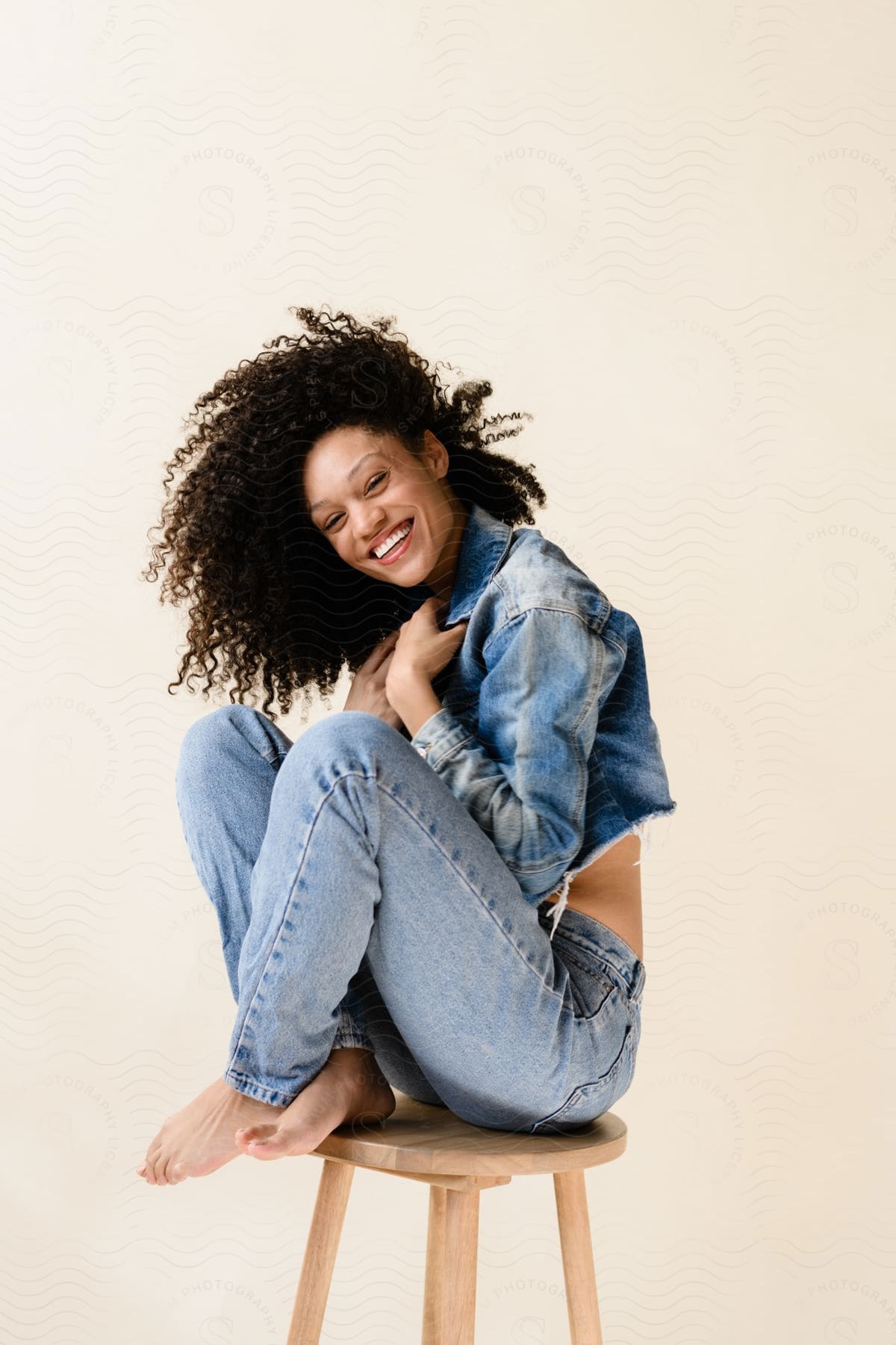 Smiling woman with curly hair sitting on a wooden stool, wearing a denim jacket and blue jeans, against a light background.