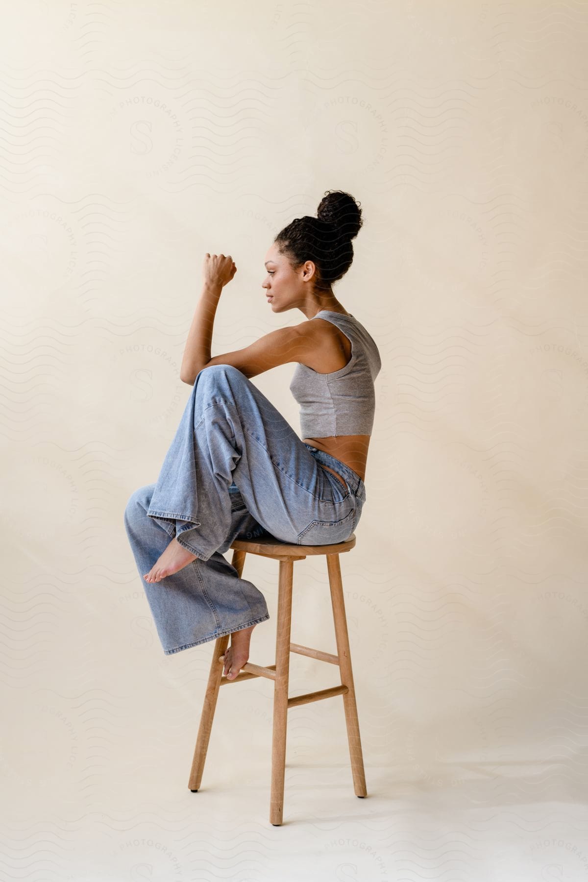 Model wearing jeans and a gray t-shirt sitting on a wooden stool on a white background in the studio